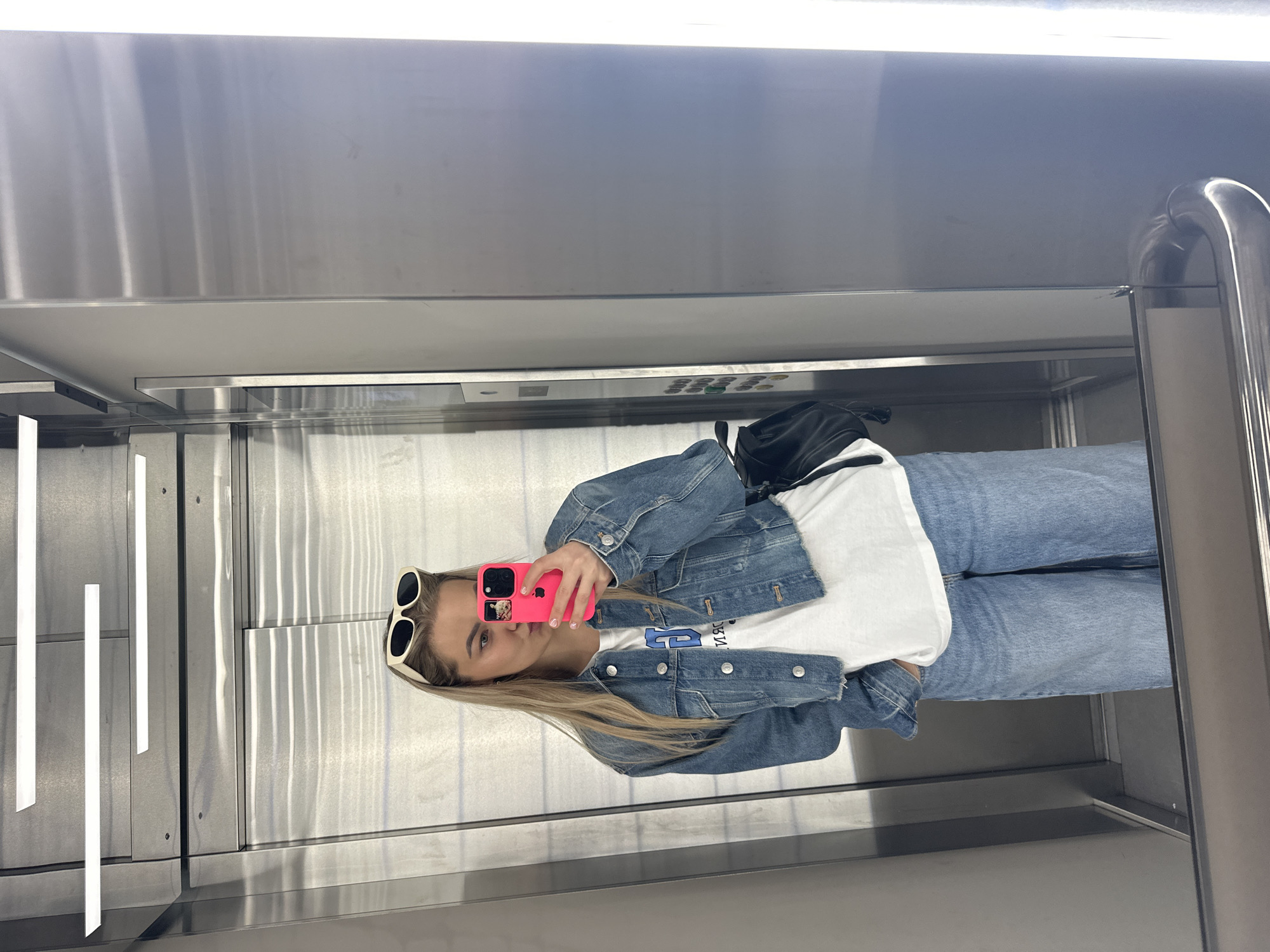 A young, blonde woman is taking a selfie in an elevator. She is wearing sunglasses, a white T-shirt, a denim jacket, and blue jeans. She holds a pink phone to take the picture. The elevator has a metallic interior, including a grab rail. The location is not apparent from the image.