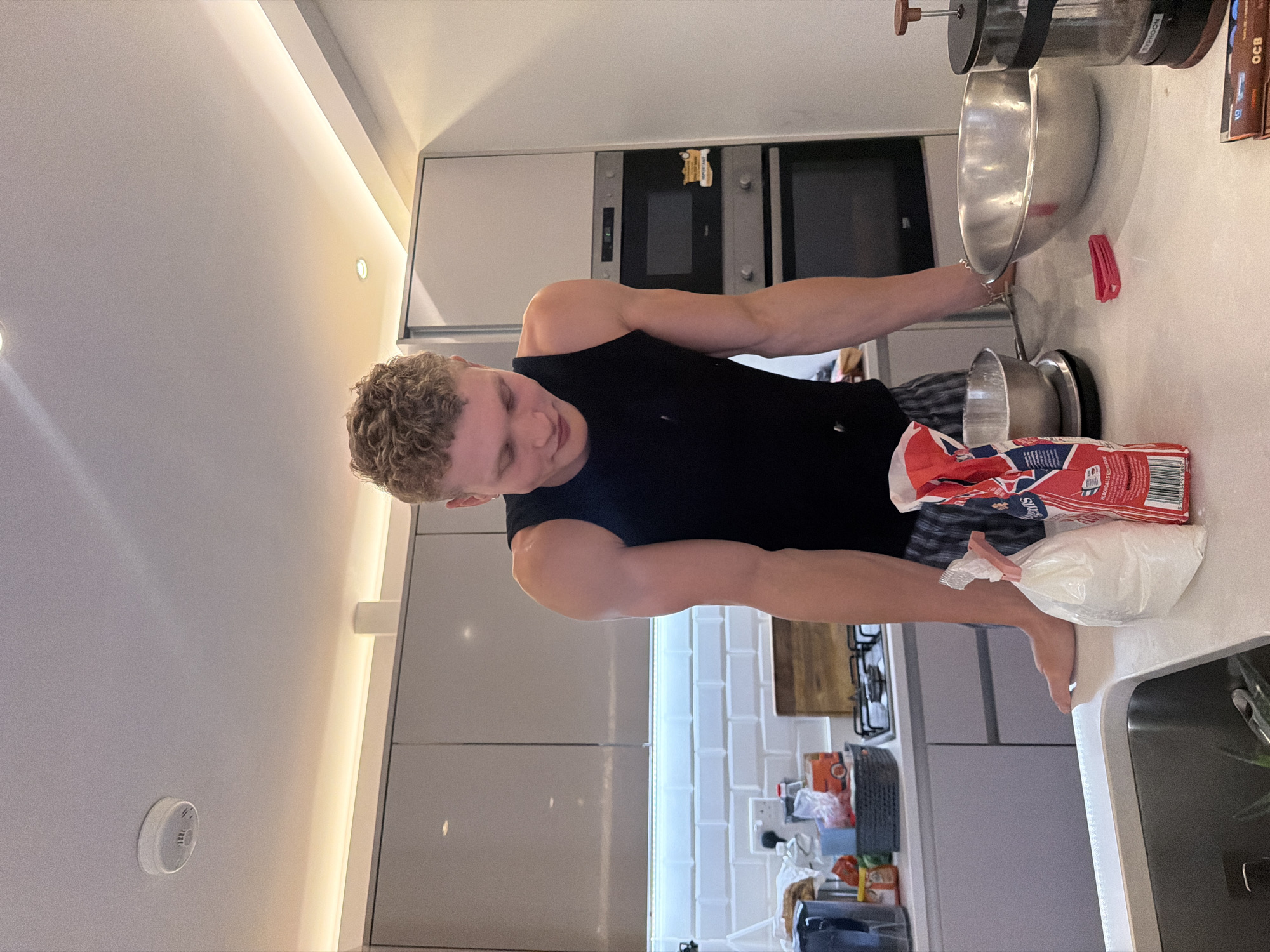 The image depicts a young man with curly blonde hair wearing a black tank top and checkered shorts, standing in a modern kitchen. He is leaning over a white countertop, looking down at a bag of white powder, likely flour, and a mixing bowl. Various kitchen items are visible, including a French press, a large metal bowl, a sieve, and a stove. The kitchen features sleek grey cabinetry, white subway tiles on the backsplash, and subtle under-cabinet lighting. The scene suggests the man is in the process of baking or preparing food. The location is a kitchen in a residential property, likely in London, United Kingdom, given the presence of a British-branded bag of flour.