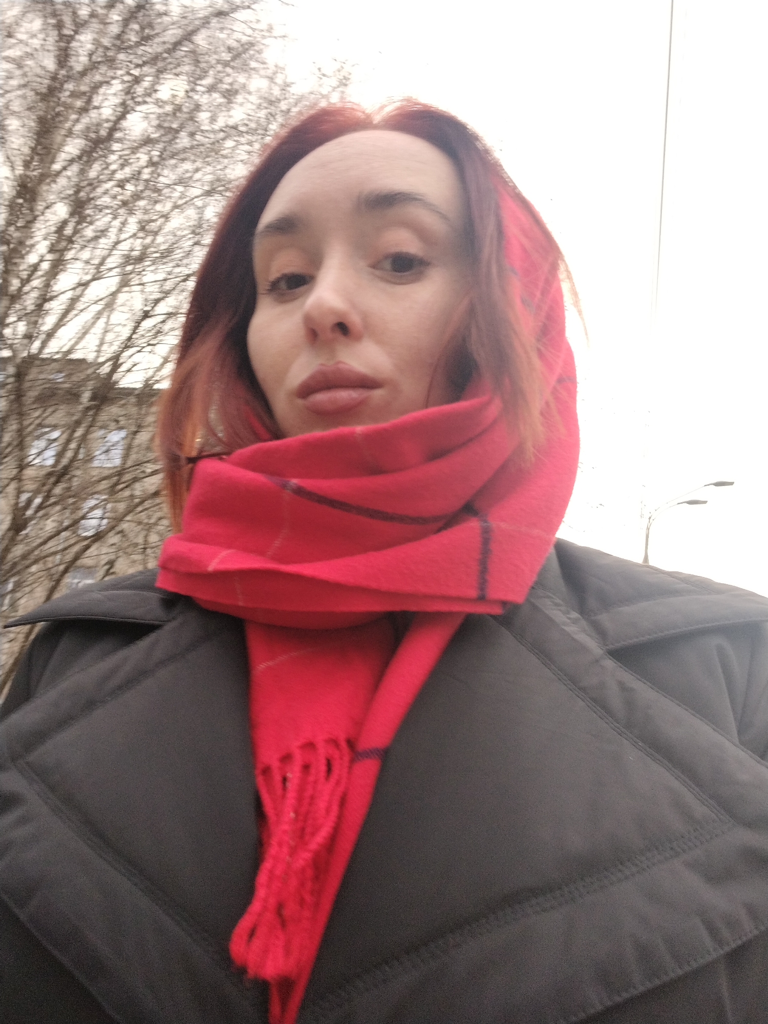 A medium close-up shot depicts a woman wearing a red plaid scarf draped over her head and shoulders, and a dark, quilted jacket. The scarf is bright red with dark plaid lines and a fringed edge. The jacket appears to be a dark grey or black, possibly a winter coat.
The background suggests an urban environment, including bare trees, a light-colored building with windows, and streetlights, the scene seems to be outdoors during the daytime.
No information is available to verify or confirm a specific location such as Izhevsk, Russia.