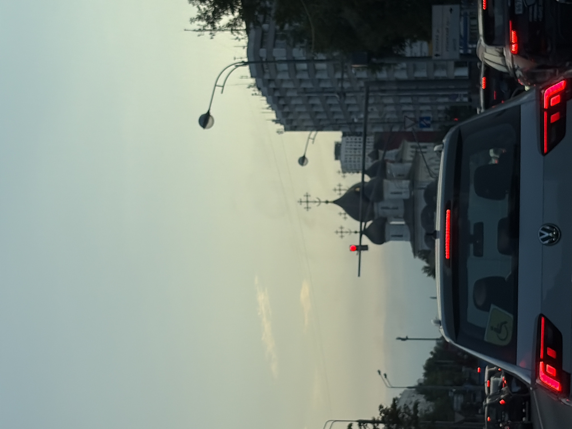 The image depicts a traffic jam in Moscow, Russia, with a view from inside a car. The foreground shows the rear of a silver car with its brake lights illuminated. A yellow sticker depicting a person in a wheelchair is visible on the rear windshield. In the middle ground, several other cars are seen in traffic. Beyond the cars, a large, white, multi-story building with many windows dominates the right side of the frame. To the left of this building, the distinct domes and crosses of an Orthodox church are visible. A traffic light with a red signal is also present. The sky above is a pale blue with a few wispy clouds, suggesting it is either early morning or late evening. The overall scene is one of urban congestion.