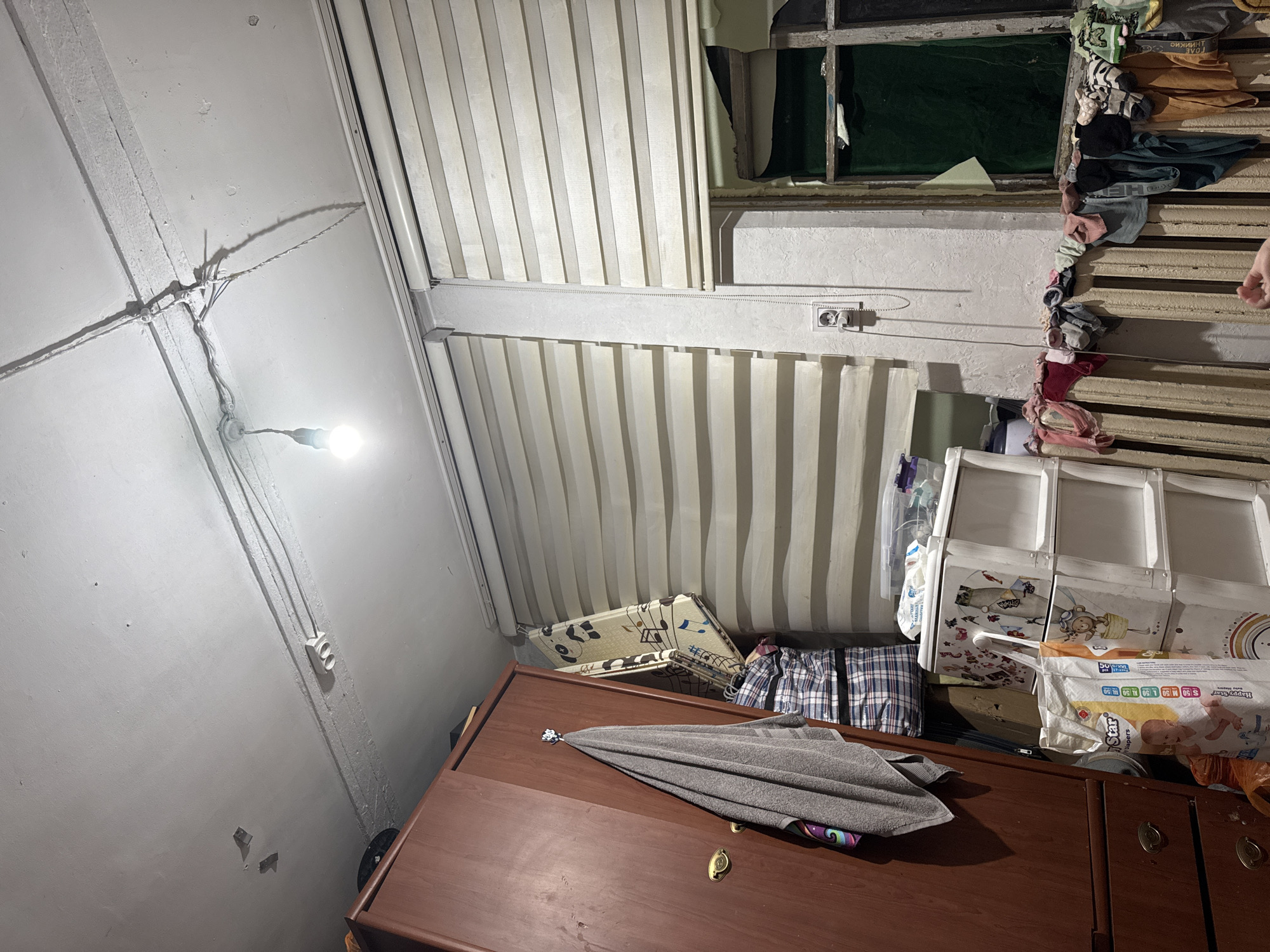 The image shows an indoor scene, likely a cluttered room. The subjects are primarily objects and the setting itself. The scene is dimly lit by a single bare light bulb hanging from the ceiling. Wires are visible, running along the white ceiling and walls. There are vertical blinds covering windows or sections of the wall. In the foreground, a dark wooden dresser or cabinet is visible, with a folded gray towel and a patterned cloth draped over it. Behind the dresser, there are stacked items, including what appear to be fabric storage cubes with animal prints and a package of diapers. Clothing items, possibly socks, are strung up along a window frame or ventilation duct on the right side of the image. The overall impression is one of disarray and utilitarian storage. There is no visible indication of the time of day or weather. The location context provided is Shamalgan, Kazakhstan, but no specific visual cues within the image confirm this. There is text visible on the diaper packaging, including "Happy Star" and size indicators like "S 50". Other text on the packaging is too small or blurry to be legible.