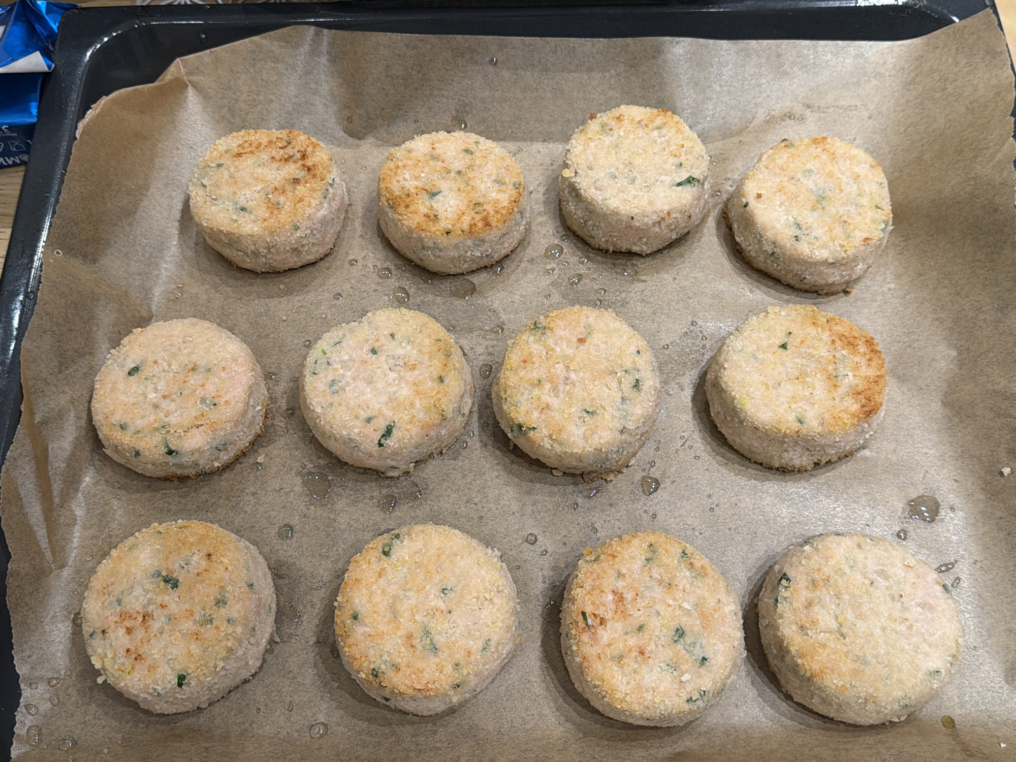 The image shows a close-up, overhead view of twelve round, breaded food patties arranged on a baking sheet lined with parchment paper. The patties are evenly spaced in a grid formation of four rows and three columns. Each patty appears to be made of a light-colored mixture, possibly fish or chicken, with visible flecks of green herbs. The breading is golden brown and appears slightly crispy, with some patties showing more browning than others. Small droplets of oil are visible on the parchment paper between some of the patties. The baking sheet itself is dark-colored and has a raised edge. In the upper right corner, a small portion of a blue and white package is visible, with some text in a foreign language. The overall scene suggests food being prepared for baking or cooking. There are no people or animals in the image. There is no indication of the location in Moscow, Russia.