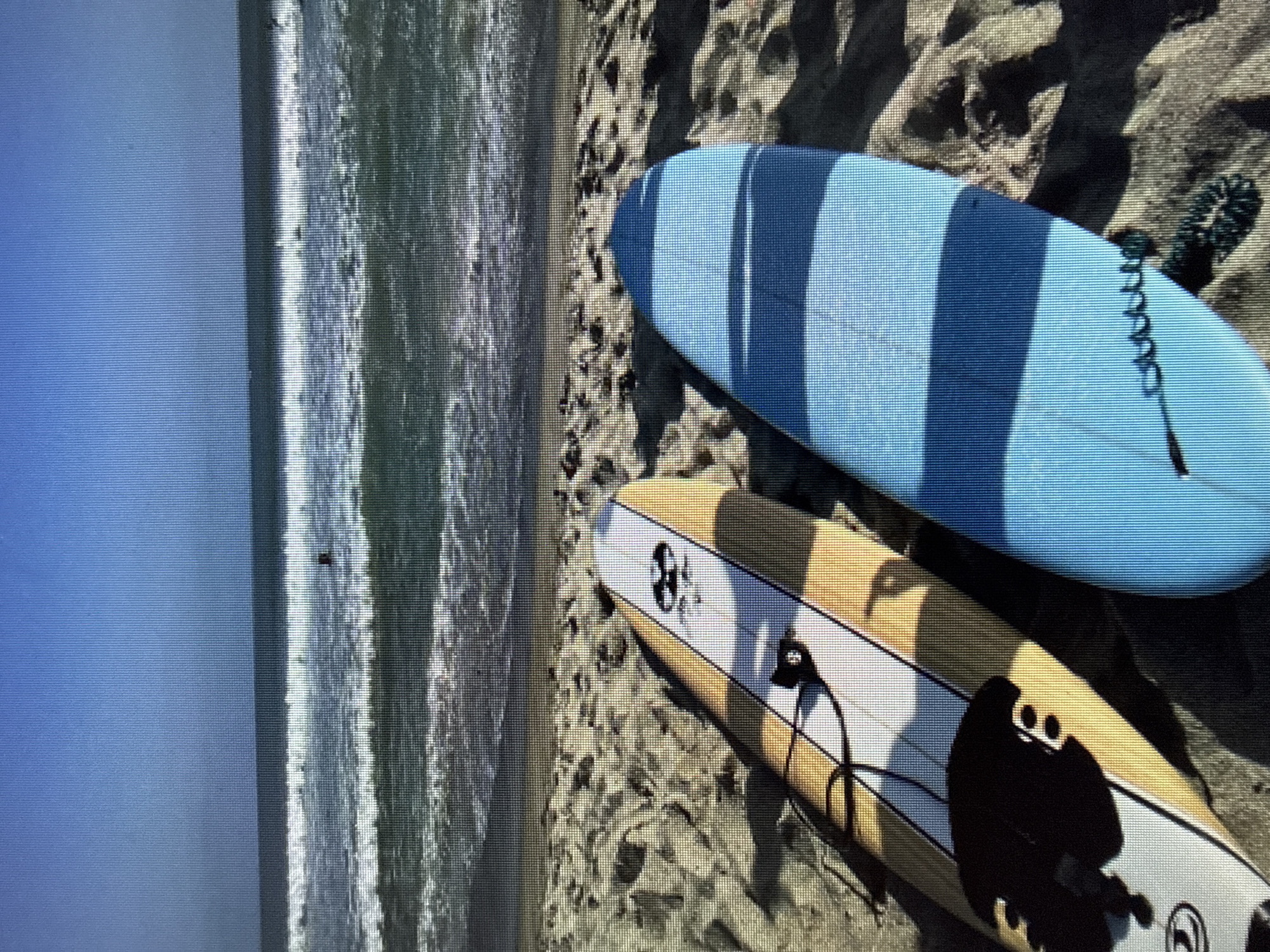 Two surfboards are on a sandy beach with the ocean in the background. The surfboard in the foreground is blue with darker blue stripes and a leash attached. The surfboard behind it is white and yellow with black markings and a leash attached. The ocean has waves and a person can be seen in the distance. The sky is clear and blue.