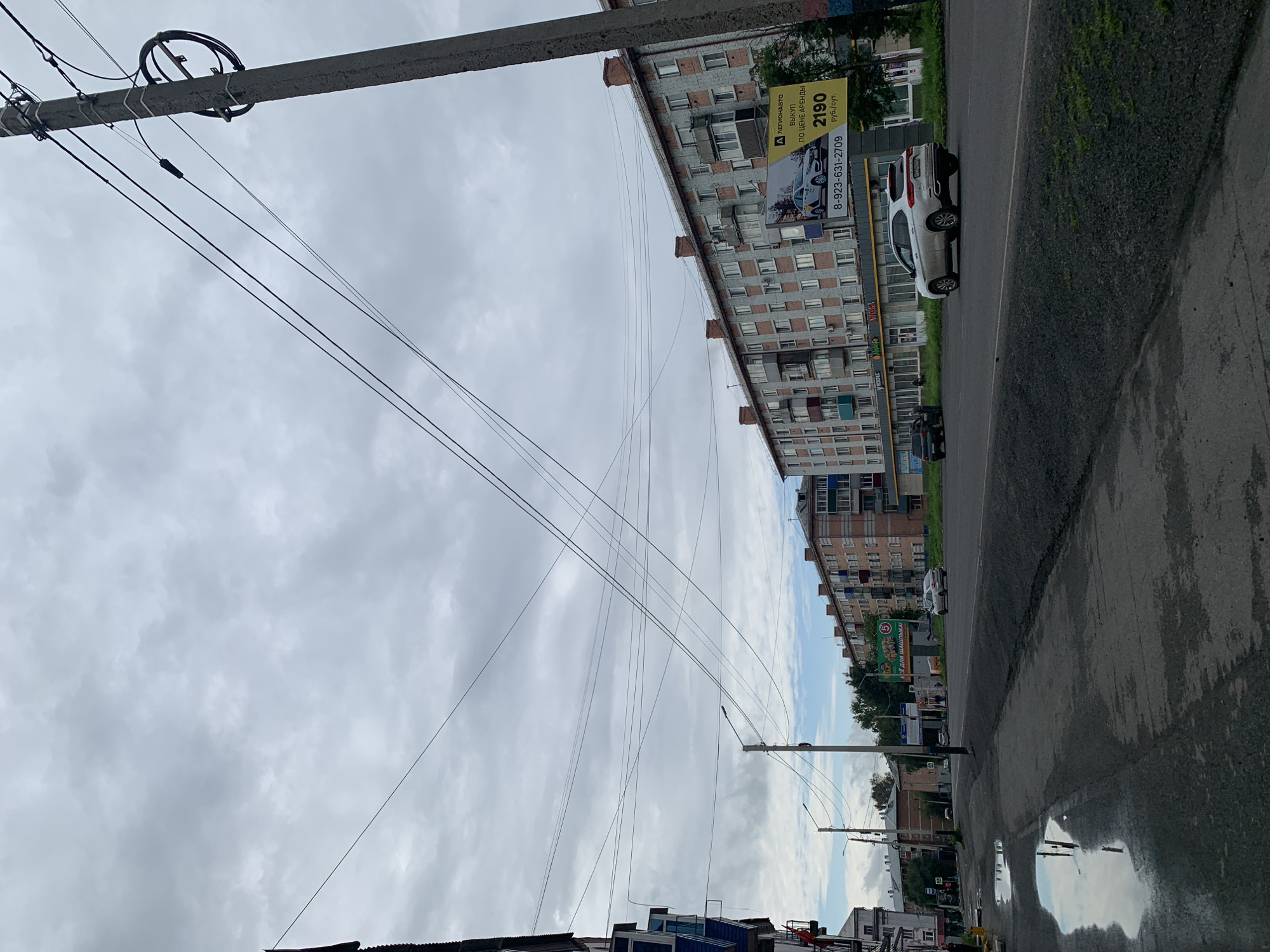 In Prokopyevsk, Russia, on a cloudy day, a street scene unfolds with multi-story apartment buildings lining a wide, asphalt road. The buildings, constructed with brick and concrete, feature numerous windows and balconies. A prominent utility pole with multiple wires stretches across the frame, dominating the sky. On the right side of the road, a large billboard displays a car advertisement with a phone number and price. Several cars are visible on the road, including a white SUV in the foreground and a black sedan in the distance. A sidewalk runs along the left side of the road, with puddles reflecting the overcast sky. The environment appears to be urban, with streetlights and some greenery visible.