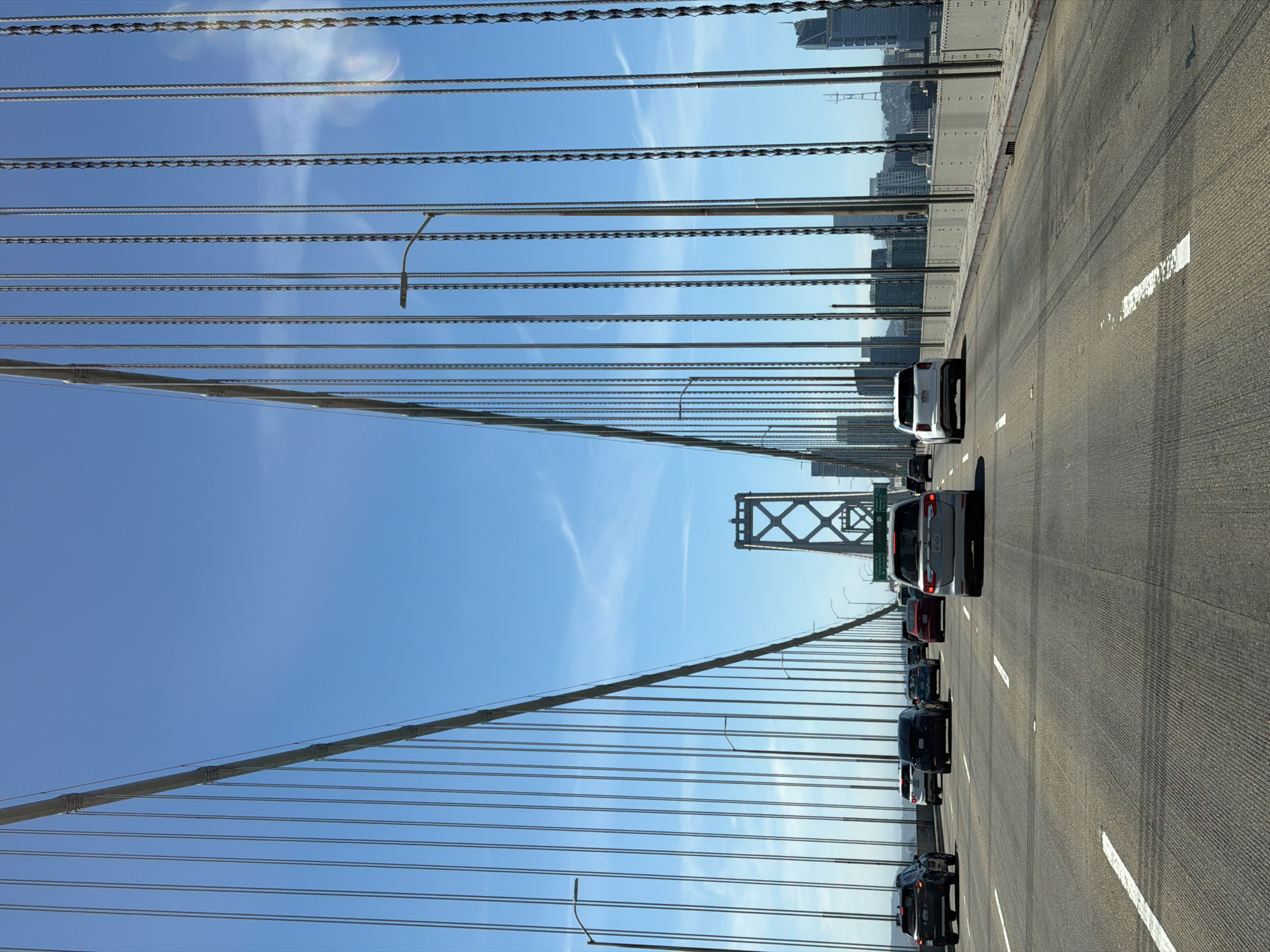 The image shows a view from a vehicle driving on a bridge in San Francisco, California, United States of America. The bridge has large suspension cables on either side, with numerous vertical cables hanging down. In the foreground, there is a multi-lane roadway with several cars driving. The cars are mostly modern SUVs and sedans. In the distance, the San Francisco skyline is visible, with tall buildings and a transmission tower. The sky is clear and blue with a few wispy clouds. The overall impression is a clear, sunny day with moderate traffic on the bridge.