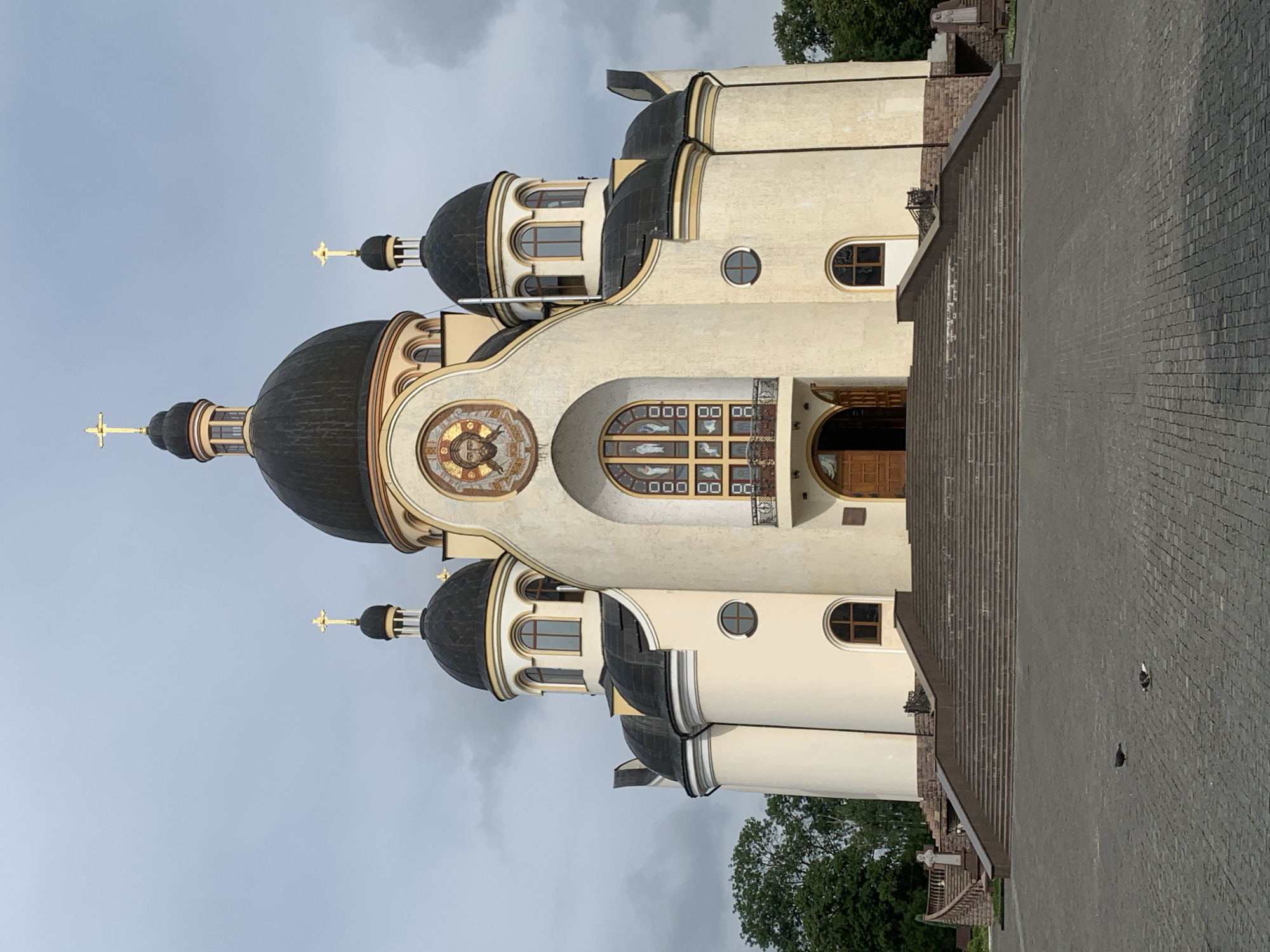 The image shows the exterior of the Church of the Holy Resurrection in Kolomyia, Ukraine, under an overcast sky. The church features a prominent central dome, flanked by smaller domes, all topped with golden crosses. The facade is cream-colored with arched windows and a mosaic of Jesus' face above the entrance. A wide set of stairs leads up to the main entrance, which is open. The ground in front of the church is paved with dark cobblestones, and two pigeons are visible on the pavement. Trees are visible on the left and right sides of the church.