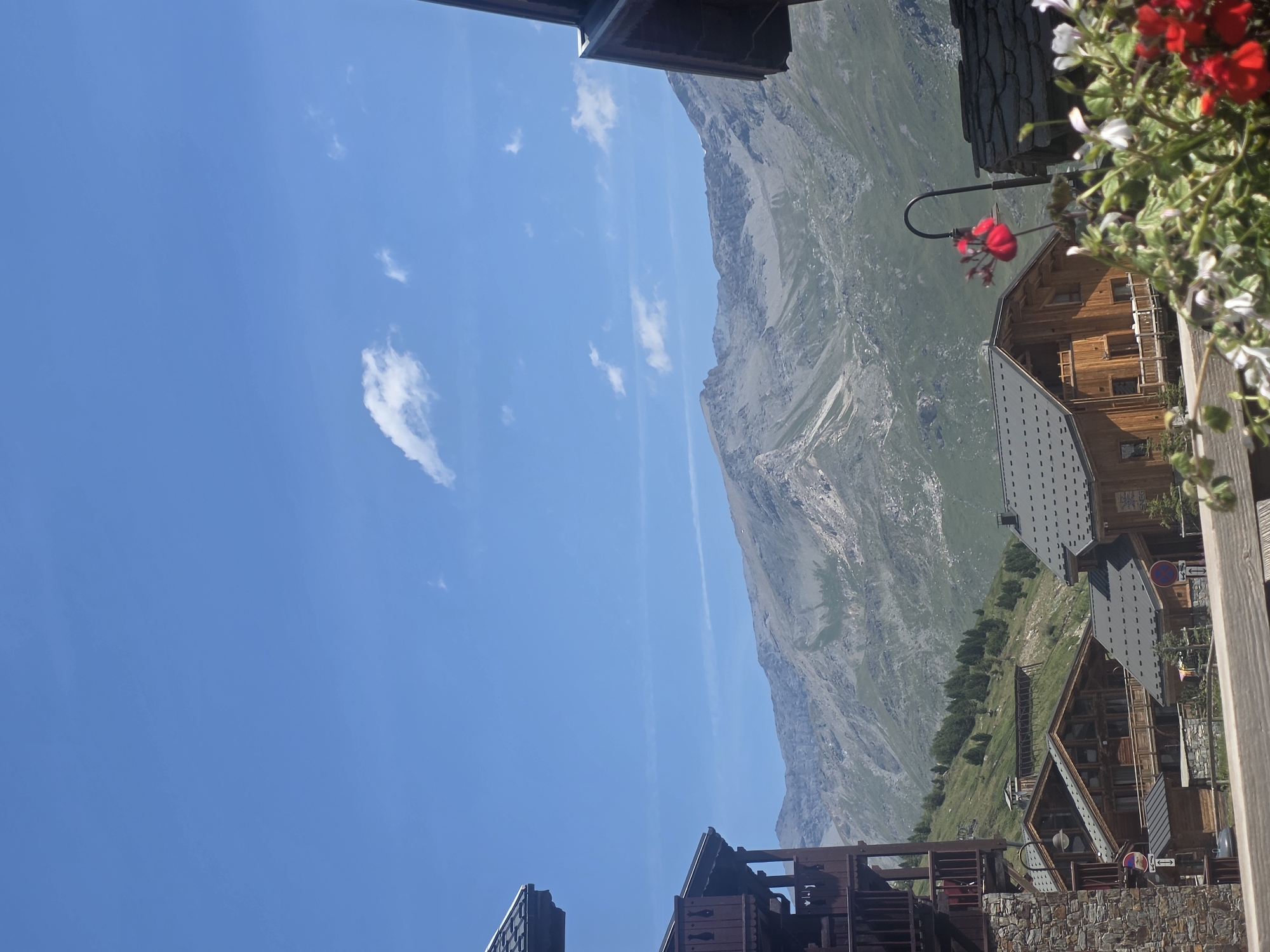 The image depicts a scenic view of a mountain village in Tignes, France, under a clear blue sky with scattered white clouds. In the foreground, traditional wooden chalets with dark grey roofs are visible, some featuring balconies and intricate woodwork. A stone building is partially visible on the left. Flowering plants with red and white blossoms add color to the scene, with a black lamppost standing among them. Behind the village, a lush green mountainside slopes upwards, dotted with trees. The background is dominated by a majestic, rugged mountain range with rocky peaks and patches of vegetation. The overall impression is one of a serene mountain resort setting.