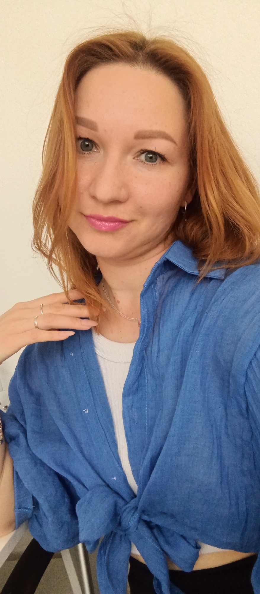 A woman with red hair and light skin is posing for a close-up selfie. She is wearing a bright blue shirt tied at the waist over a white tank top. Her left hand is resting on her neck, and she is wearing two rings on her left hand. A delicate silver necklace is visible around her neck. The background is a plain white wall.
The location is Unknown, Unknown.