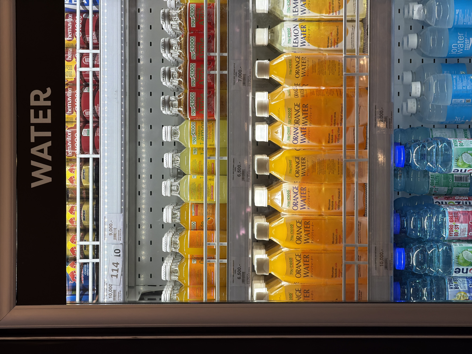 This vertical shot captures an organized refrigerated display case in a retail setting, likely a convenience store or supermarket, in North Kuta, Indonesia.
The display features a variety of beverages arranged on shelves. On the far left, a prominent vertical "WATER" sign is visible. Beneath it are several rows of canned energy drinks, including red cans of "hemaviton," gold cans of "KRATINGDAENG SUPER ENERGY DRINK," and red cans of "RED BULL CLASSIC ENERGY DRINK," some marked with "HALAL." Price tags of "10,000" and "9,000" are seen in this section.
The middle and main section of the cooler is stocked with "YOU-C1000" brand vitamin and isotonic drinks in plastic bottles. Flavors include "VITAMIN LEMON," "VITAMIN APPLE," and "ORANGE WATER," with corresponding Japanese script ("レモンウォーター," "オレンジウォーター"). Labels mention "Minuman Isotonik unik dengan 1000 mg Vitamin C" (Unique Isotonic drink with 1000 mg Vitamin C), "Isi bersih: 500 ml" (Net content: 500 ml), and "Healthy Inside Fresh Outside." Prices such as "6,500.-" for the 140ml vitamin drinks and "7,000.-" for the apple flavor are visible.
The rightmost section contains various brands of bottled water and isotonic drinks, including "ion water," "POCARI SWEAT," and "HYDRO BOOST," which features "HYDRO GREAT BOOST TASTE" and "one." A price tag of "10,000.-" is also visible in this area. The products are neatly aligned, and the cooler is well-lit by internal lights.