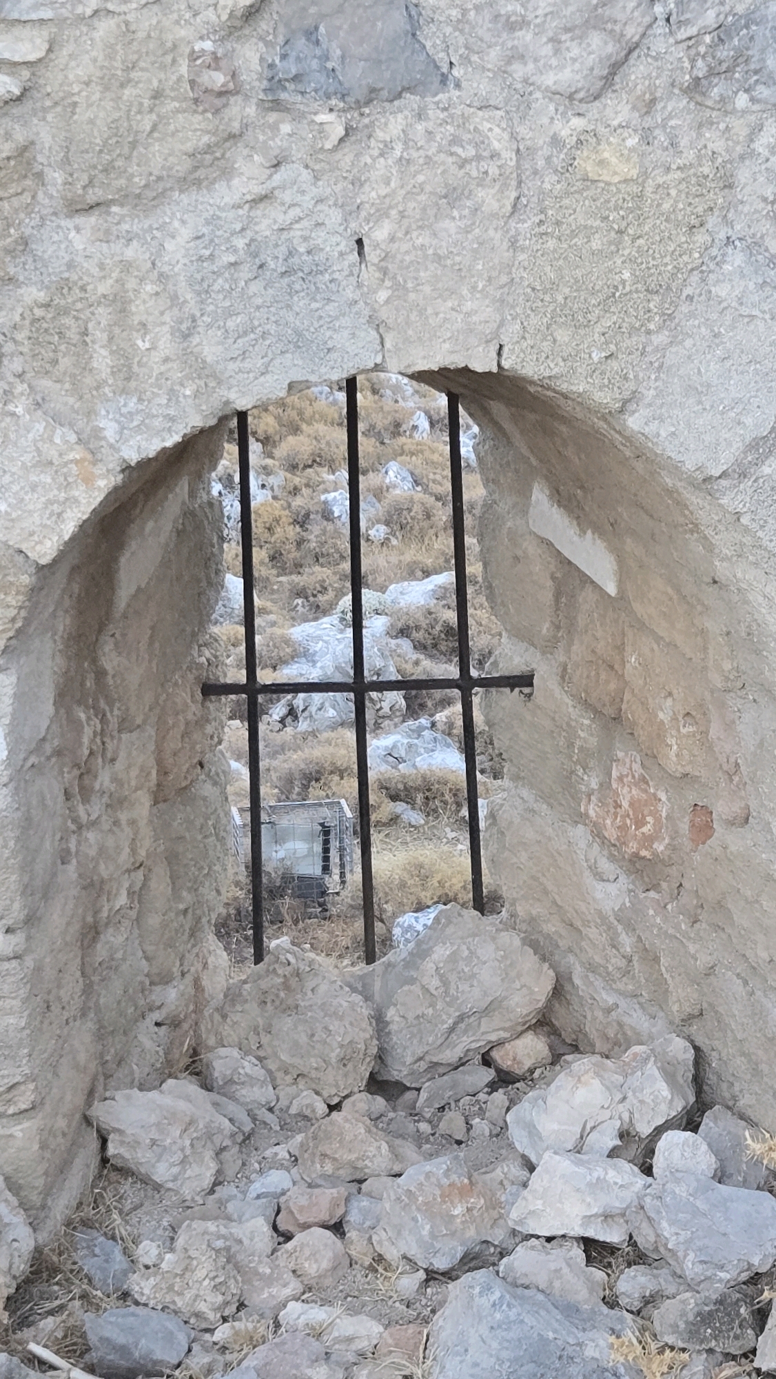 The image depicts an arched opening in a weathered stone wall, secured by a metal grate. Through the grate, a dry, rocky, and shrub-covered hillside is visible. In the distance, partially obscured by the grate, is a metal structure that appears to be a cage or enclosure. The foreground is filled with a jumble of rocks and dry vegetation. The scene is likely located in the vicinity of Archangelos, Greece, given the characteristic stone construction and arid landscape common to the region. There are no people or discernible actions taking place.