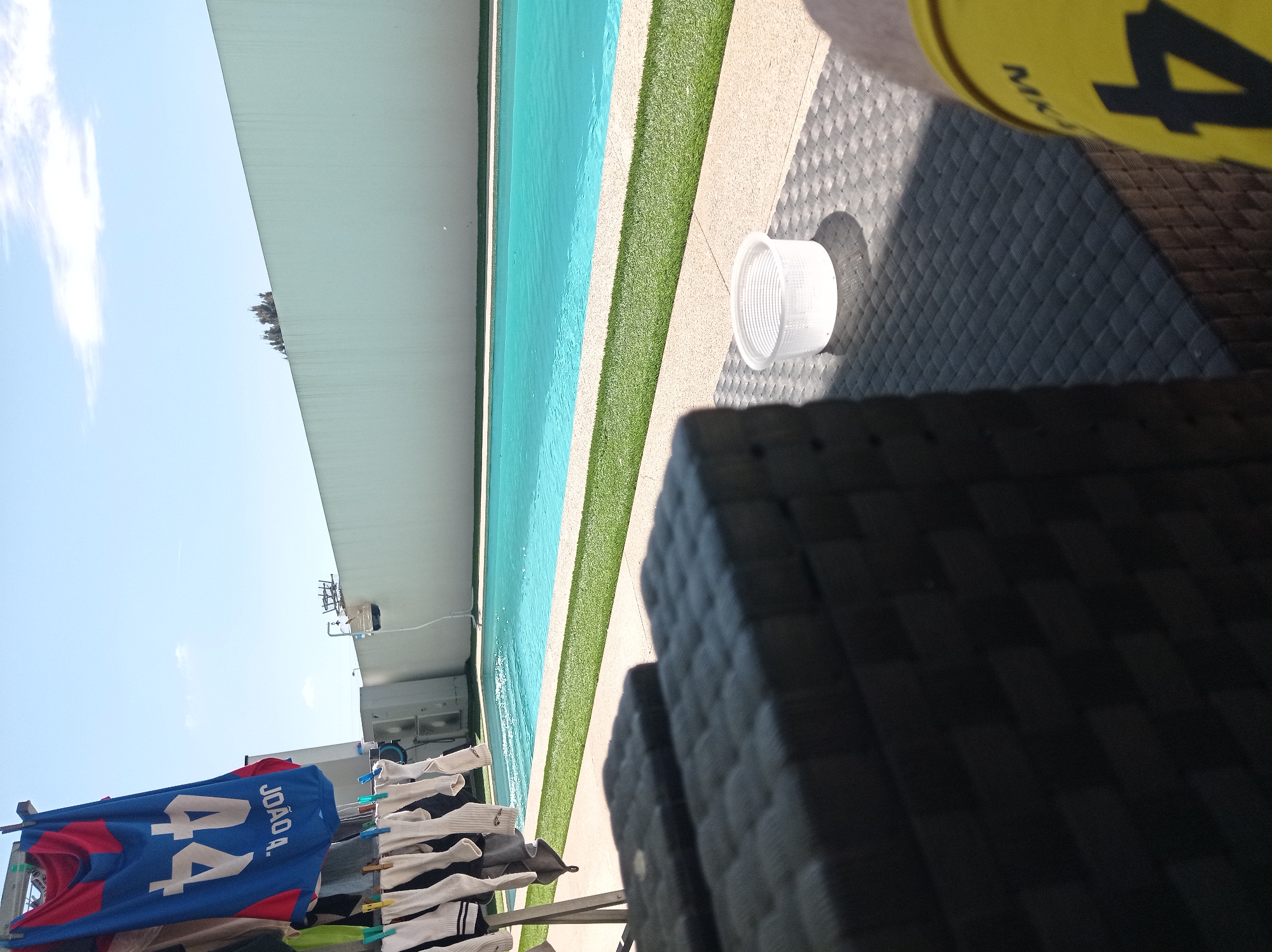 The scene depicts an outdoor residential area in Felgueiras, Portugal, under bright daylight. The perspective is from a low vantage point, likely from a seated position.
In the immediate foreground, a dark-colored, woven outdoor furniture surface (possibly a tabletop or armrest) is visible, partially obscuring the lower-left and central portions of the view. To the left, a metallic clothes drying rack stands, displaying various items of laundry. Notable items include a blue and red sports jersey with the upside-down text "JOÃO A." and the number "44" in white, along with multiple pairs of white socks and other light-colored garments attached with clothespins. In the lower-right quadrant, a portion of a human leg is visible, clad in yellow shorts or swimwear featuring a black number "4" and partial text that appears to be "MIK".
The midground features a rectangular swimming pool filled with clear blue water, reflecting the sunlight. The pool is bordered by light-colored paving, beyond which a strip of green artificial turf runs parallel to the pool's edge. A small, white, cylindrical plastic basket with a ribbed exterior rests on the dark woven surface, casting a shadow to its right. Beyond the pool, against a white exterior building wall, two outdoor air conditioning units are mounted. A thin pole with a small basket-like attachment is visible near the far end of the pool.
The background is dominated by a tall, plain, light-colored (possibly off-white or light grey) perimeter wall that spans horizontally across the upper portion of the scene. Above this wall, the tops of several trees or dense foliage are visible. The sky is clear blue with sparse white clouds, and a faint aircraft contrail is discernible in the upper sky.