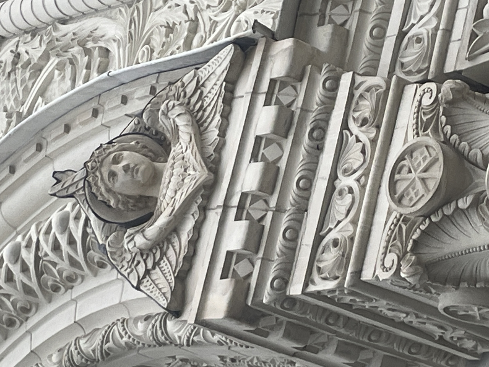 The image shows a close-up of intricate stone carvings on the facade of a building in Kronstadt, Russia. The primary subject is a winged angelic figure's head and upper torso, carved in relief. The angel wears a halo or crown and has feathered wings spread behind its head. Below the angel, the architectural details include decorative friezes with geometric patterns, scrollwork, and acanthus leaf motifs. The stone appears weathered, suggesting the building is historic. The lighting is diffused, indicating an overcast sky or the building is in shadow. The perspective is from below, looking upwards at the architectural elements.