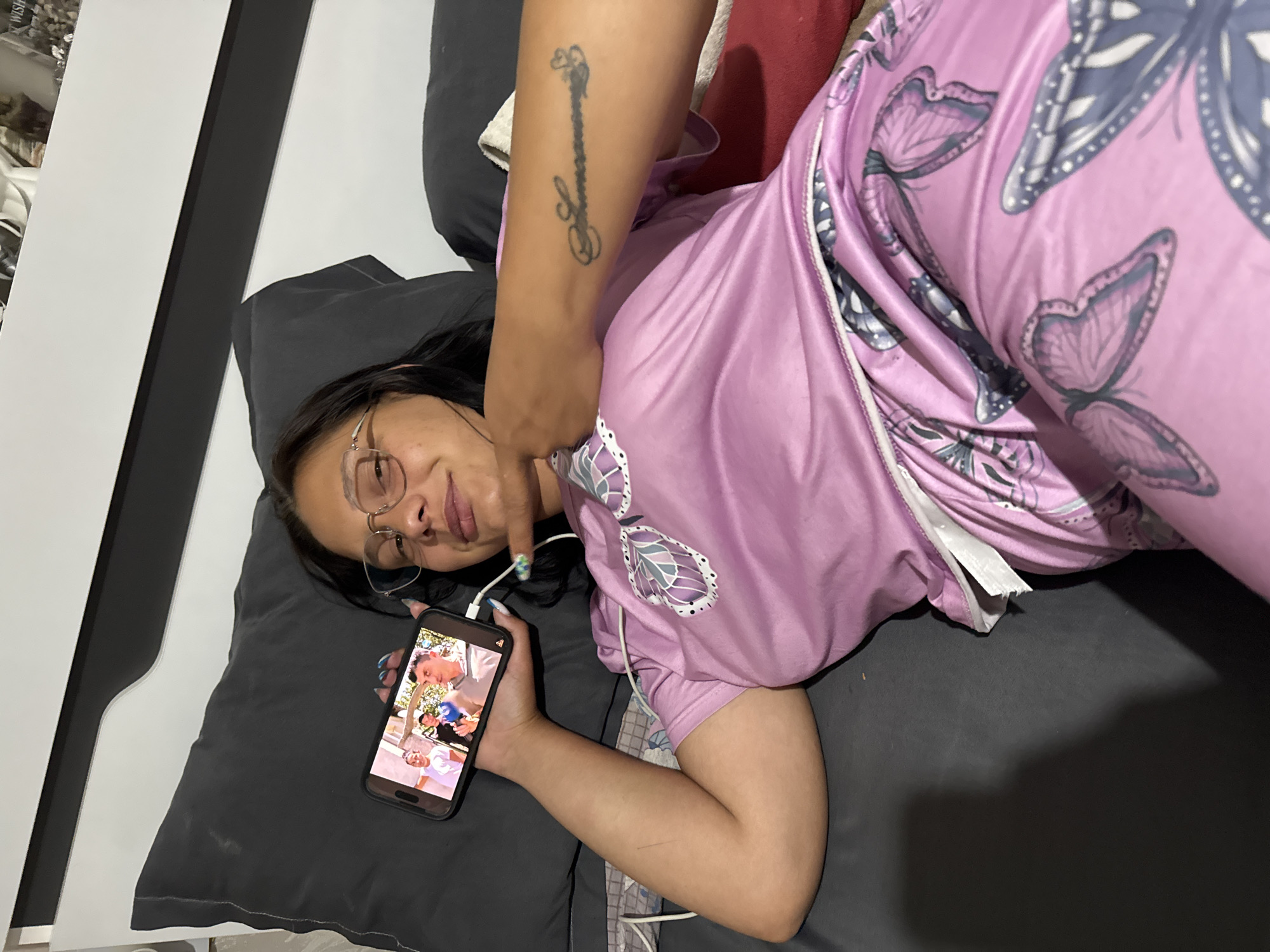 A woman is lying on a gray bed or couch, propped up by gray pillows. She is wearing glasses and a pink satin pajama set patterned with gray butterflies. She is holding a black smartphone in her left hand, displaying an image or video of people. Someone's tattooed arm is draped over her. A white object is visible behind her, and a glimpse of a red item can be seen in the background. The location is unlikely to be identifiable from the image without further information.