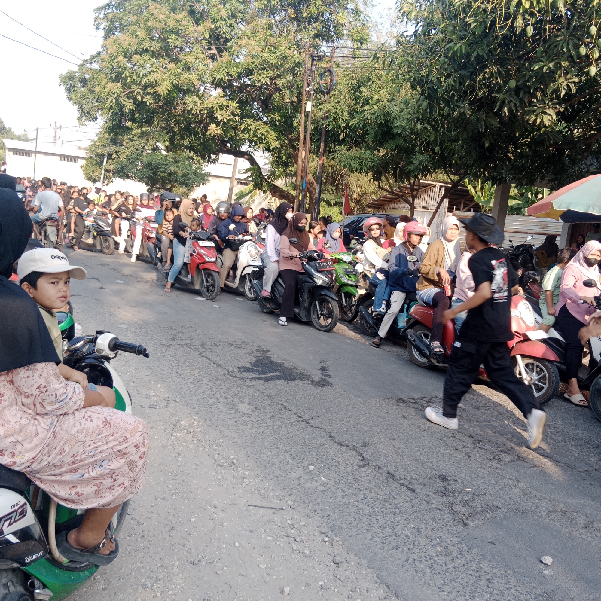 The image depicts a street scene in Tegalrejo, Indonesia, with a large group of people gathered, primarily on motorbikes. The setting appears to be a sunny day, with trees lining the street. The road is paved and shows signs of wear.
In the foreground, a young boy wearing a white baseball cap and a light-colored shirt is seated on a green and white motorbike, looking to his right. He is partially obscured by a person in a black headscarf and a pink floral dress. The motorbike has branding that includes "Mio" and "Yulfi.fi".
The middle ground is filled with a dense crowd of people, many of whom are on motorbikes. The motorbikes are varied in color, including red, black, white, and green. The people are dressed in casual attire, with many women wearing headscarves (hijabs). Some individuals are wearing helmets. There are adults and children among the crowd. The motorbikes are lined up, suggesting a traffic jam or a gathering.
In the background, more trees and some buildings are visible. A red flag is planted on the right side of the street. A partially visible umbrella or canopy with red, yellow, and green sections is also present. The sky is bright, indicating daylight. The overall atmosphere suggests a busy and crowded urban environment.