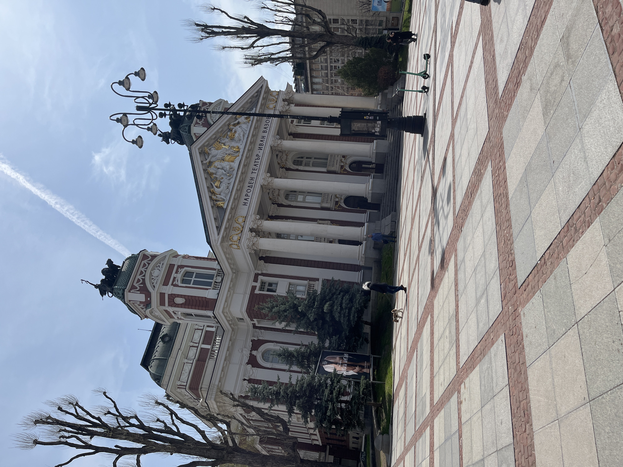 The image depicts the Ivan Vazov National Theatre in Sofia, Bulgaria, as seen from a ground-level perspective. The building's facade is predominantly white with red brick accents and features classical architectural elements such as columns and a triangular pediment adorned with sculptural details. Text is visible on the building's front. A lamppost stands prominently in the foreground, adding verticality to the composition. Bare-branched trees flank the theater, suggesting it might be late autumn or winter. In the background, other buildings are visible, indicating the urban setting. The foreground is dominated by a pedestrian plaza paved with square tiles, some of which are reddish-brown. A woman walking a dog is on the right side of the image. Two scooters are parked at the top of the image on the right.