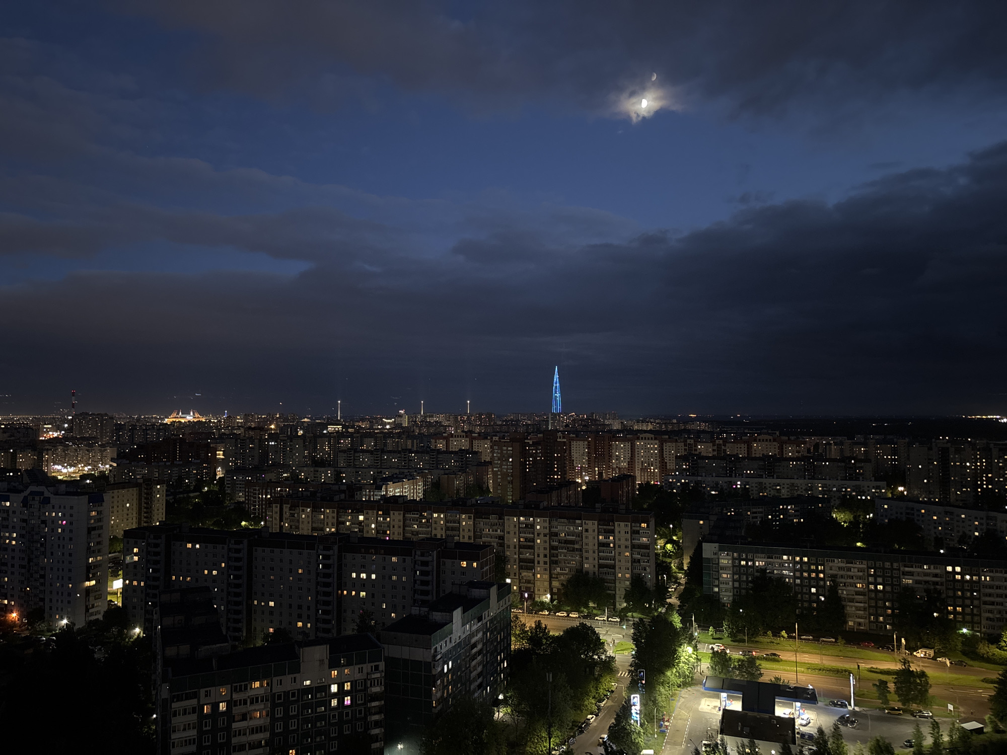 The image shows a nighttime cityscape of Saint Petersburg, Russia. In the distance, the illuminated Lakhta Center skyscraper is visible. A crescent moon is present in the sky, partially obscured by clouds. The foreground is dominated by residential buildings with illuminated windows. Streetlights are on, and cars are parked along the street. A gas station is also visible.