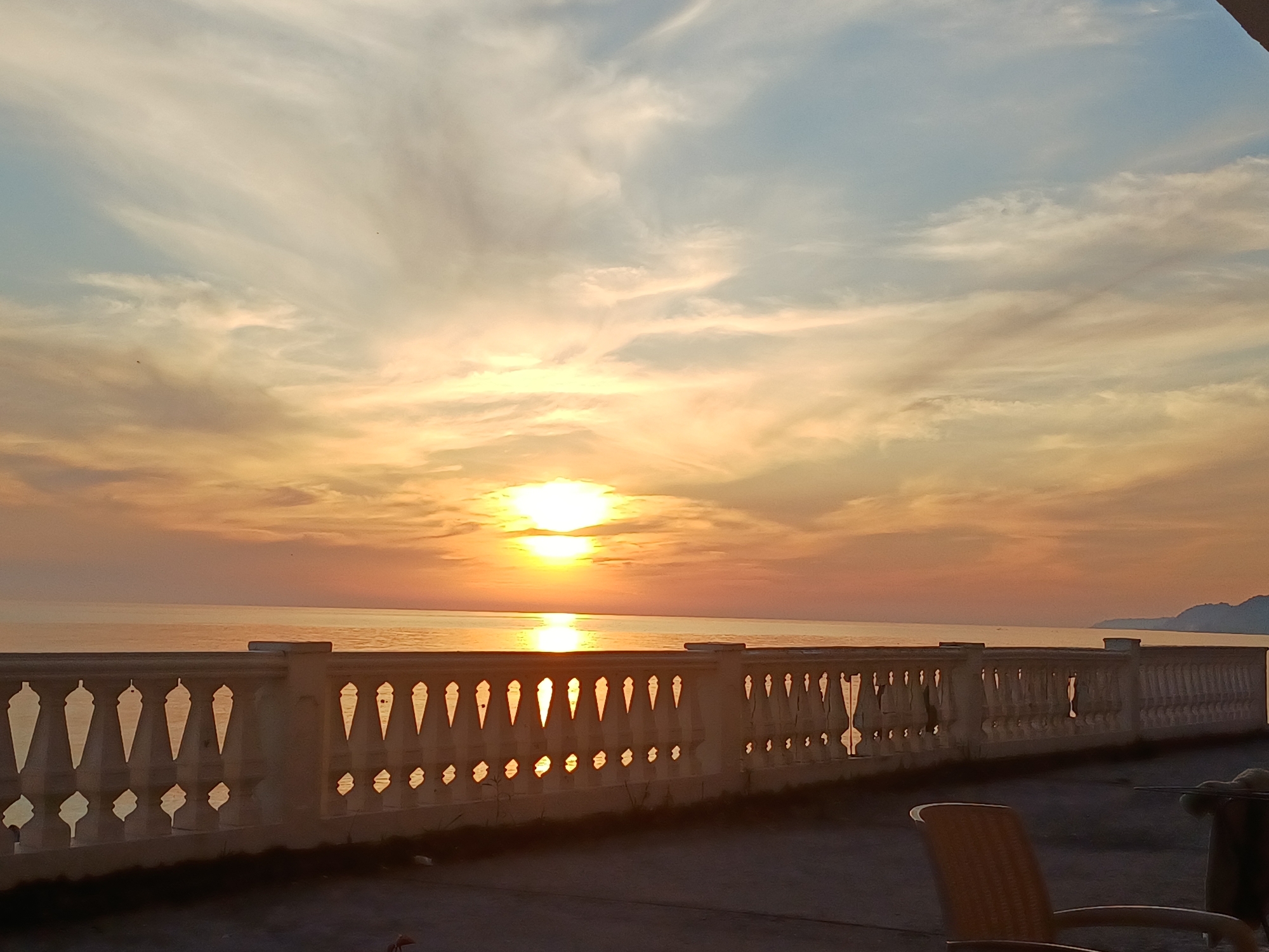 The image displays a sunset scene over a body of water, likely the Black Sea, viewed from a terrace with a decorative balustrade in Sochi, Russia.
The sky in the upper half of the frame presents a gradient of colors, transitioning from light blue at the top left to warm orange and golden yellow tones towards the horizon. Wispy, elongated clouds (cirrus) and more diffuse, patchy clouds (stratocumulus) are illuminated by the setting sun, enhancing the warm color palette. The sun itself, a brilliant, nearly circular source of light, is positioned centrally on the horizon, partially veiled by clouds, creating a strong radiant glow. Two small, dark, bird-like specks are visible against the sky in the upper left quadrant.
The mid-ground is dominated by a calm body of water, stretching from the foreground balustrade to the distant horizon. The water's surface reflects the vivid orange and yellow light from the sky, with a prominent, elongated, shimmering reflection of the sun extending directly towards the viewer.
In the lower foreground, a substantial, light-colored concrete balustrade or railing spans across the scene. This balustrade features a series of intricately designed geometric cut-outs, resembling inverted teardrops or stylized keyholes in an upper row, and smaller, similarly shaped openings in a lower row. The bright sunlight passes through these apertures, casting reflections onto the water and creating defined light patterns on the balustrade itself. Below the railing is a dark, flat surface, consistent with a paved terrace or balcony floor, with sparse patches of dried vegetation visible along the base of the balustrade. To the far right in the foreground, part of a light brown, possibly woven or plastic, outdoor chair with an armrest is visible, along with a dark, horizontal rod-like object that extends from the right edge of the frame.
In the distant background, on the far right along the horizon line, a faint, dark silhouette of a low-lying landmass or mountain range is discernible, confirming a coastal environment characteristic of Sochi.