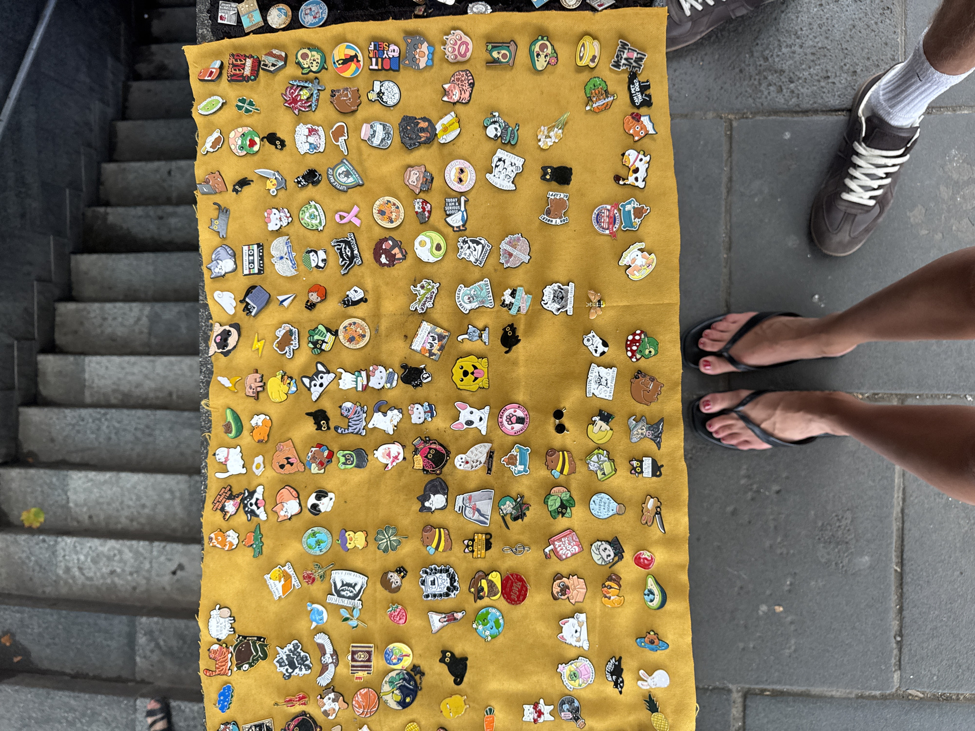 The image shows an overhead view of an outdoor stall displaying numerous enamel pins on a yellow fabric. The stall is set up on a stone-paved ground, with grey stone steps visible in the background. Two people's feet are visible in the foreground, one wearing black flip-flops and the other wearing dark brown sneakers with white laces and white socks. The pins are assorted and depict various subjects including animals (cats, dogs, sheep, birds, foxes, rabbits), food items (avocado, strawberry, pineapple), objects (lightbulb, passport, books, violin), and abstract designs (yin-yang, colorful circles, medical symbols, hearts, pink ribbon). Some pins feature text, such as "GOOD FRIENDS," "STAY TRASHY," "NEVER 10000," and "CAN I PET THAT DOG?". The stall appears to be located in a public area with pedestrian traffic. While the image does not contain explicit markers of Tbilisi, Georgia, such outdoor stalls selling accessories are common in pedestrian zones and markets in cities like Tbilisi, particularly in areas with historical significance or tourist appeal. The general urban setting with stone paving and steps is consistent with the architectural style found in Tbilisi.