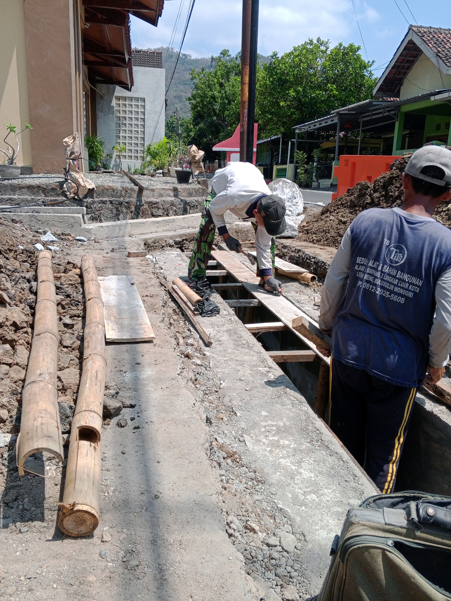 In Giritirto, Indonesia, a construction site is depicted under a clear sky. Two men are present, actively engaged in labor. One man, wearing a white long-sleeved shirt, camouflage pants, and a black cap, is positioned in the center of the frame. He is stooped over, holding a tool in his right hand and interacting with a wooden plank laid across a trench. His left hand is also near the plank.
Another man, seen from the back, is wearing a blue long-sleeved shirt with text and a gray baseball cap. He stands to the right, his body angled towards the trench, his hands at his sides. His shirt displays a logo and text, including "SEDIA: ALAT & BAHAN BANGUNAN" and a phone number.
The environment is characterized by a partially dug trench, with exposed soil and concrete structures. Two large bamboo poles are laid horizontally on the ground to the left of the trench. Construction materials and debris are scattered around the site. In the foreground, a dark green duffel bag is partially open, resting on the ground. The background reveals houses with tiled roofs and lush green trees, suggesting a semi-urban or suburban setting. The overall scene captures a moment of ongoing construction work in a residential area.