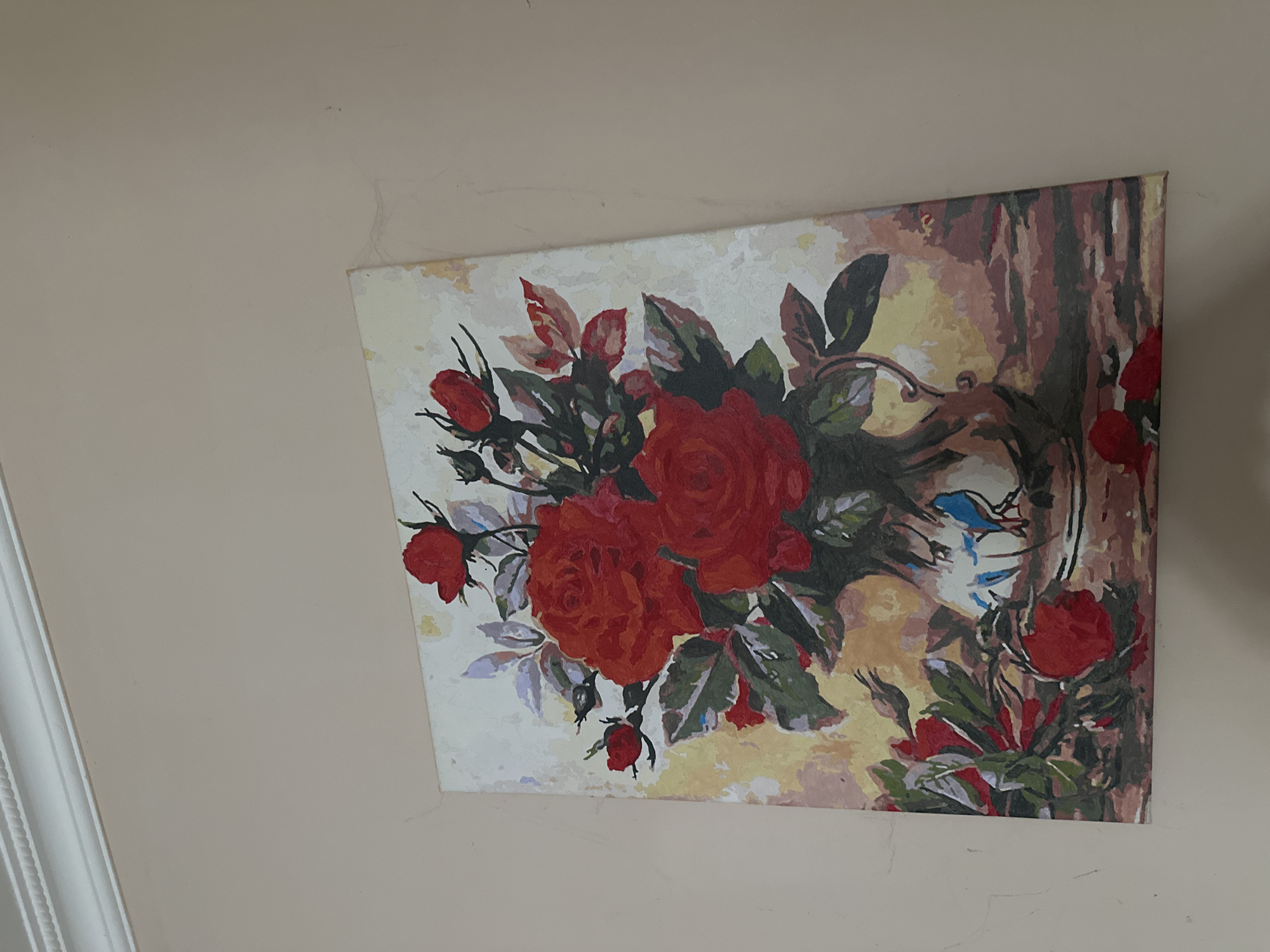 The image shows a painting of a vase of red roses. The painting is hanging on a light-colored wall, and there is decorative molding at the top of the wall. The painting appears to be a still life, with the roses arranged in a vase on a table. The background of the painting is a soft, muted yellow and brown. There are several red roses, some in full bloom and others as buds. There are also green leaves and stems. A blue bird is depicted on the vase. The painting has a textured appearance, suggesting it might be a print or a painting by numbers. The scene is static and does not depict any action or interaction. The location is not specified in the image, and therefore cannot be determined.