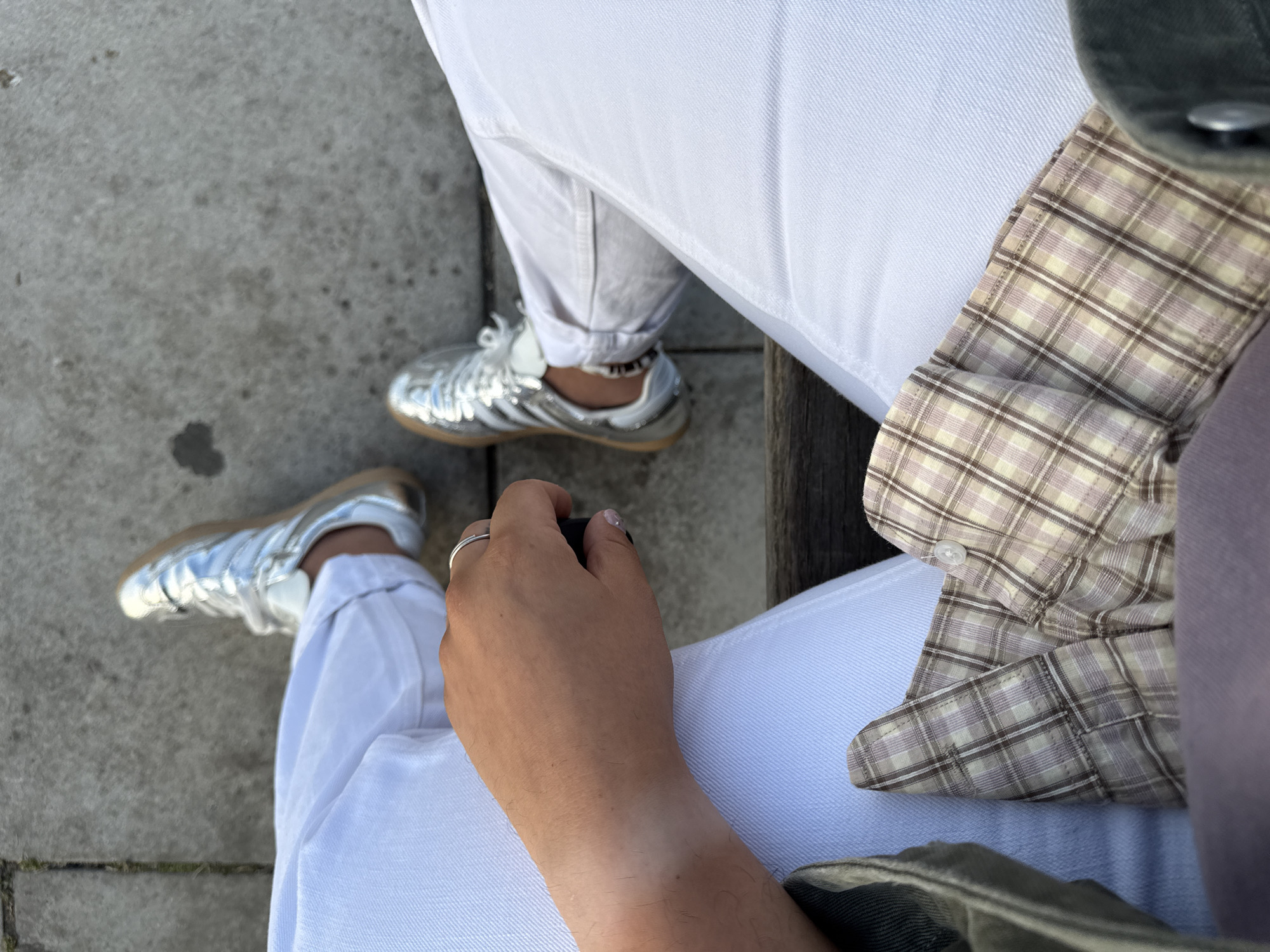 This is a close-up, eye-level shot of a person's legs and feet, seated on a wooden bench outdoors, with a concrete pavement in the background. The person is wearing white jeans, a plaid button-up shirt with muted colors of cream, light brown, and pink, and silver athletic shoes with tan soles. The person's right hand is visible, resting on their leg, and they are wearing a silver ring on their left hand. The scene appears to be in Vila Nova de Gaia, Portugal, possibly near the Douro River, given the typical pedestrian areas found there.