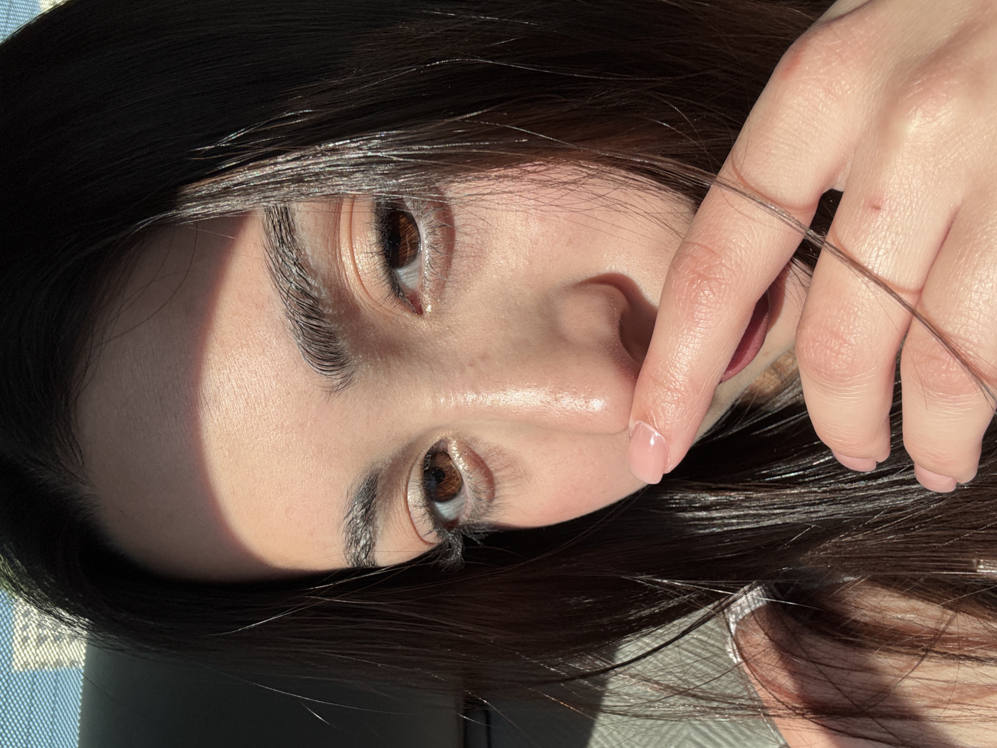 A close-up, rotated portrait shows a person's face, angled to the right and slightly tilted upward. The person has dark, glossy hair, brown eyes, and fair skin. A hand is positioned near the face, with a single hair strand crossing between the fingers. The lighting is bright, casting shadows across half of the face. Based on the available information, it is not possible to determine if the photo was taken in Yerevan, Armenia.