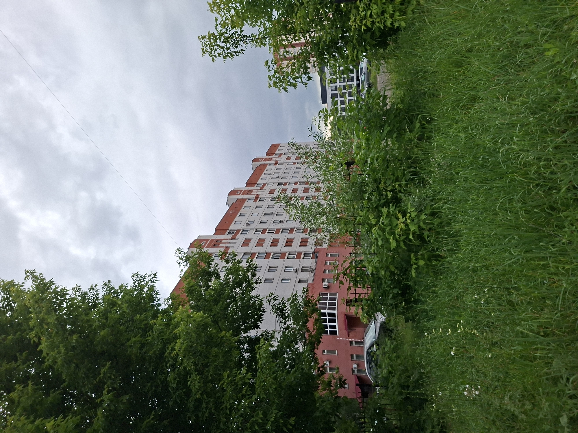 The image shows a residential building in Kazan, Russia. The building is tall and multi-story, with a red and white facade. There is a large tree in the foreground on the left, with green foliage. A grassy area with some wildflowers is in the foreground. A dark-colored car is parked in front of the building, behind a black metal fence. Other trees and greenery are visible. The sky is overcast and gray. The scene depicts an outdoor environment with a residential structure, vegetation, and a vehicle.