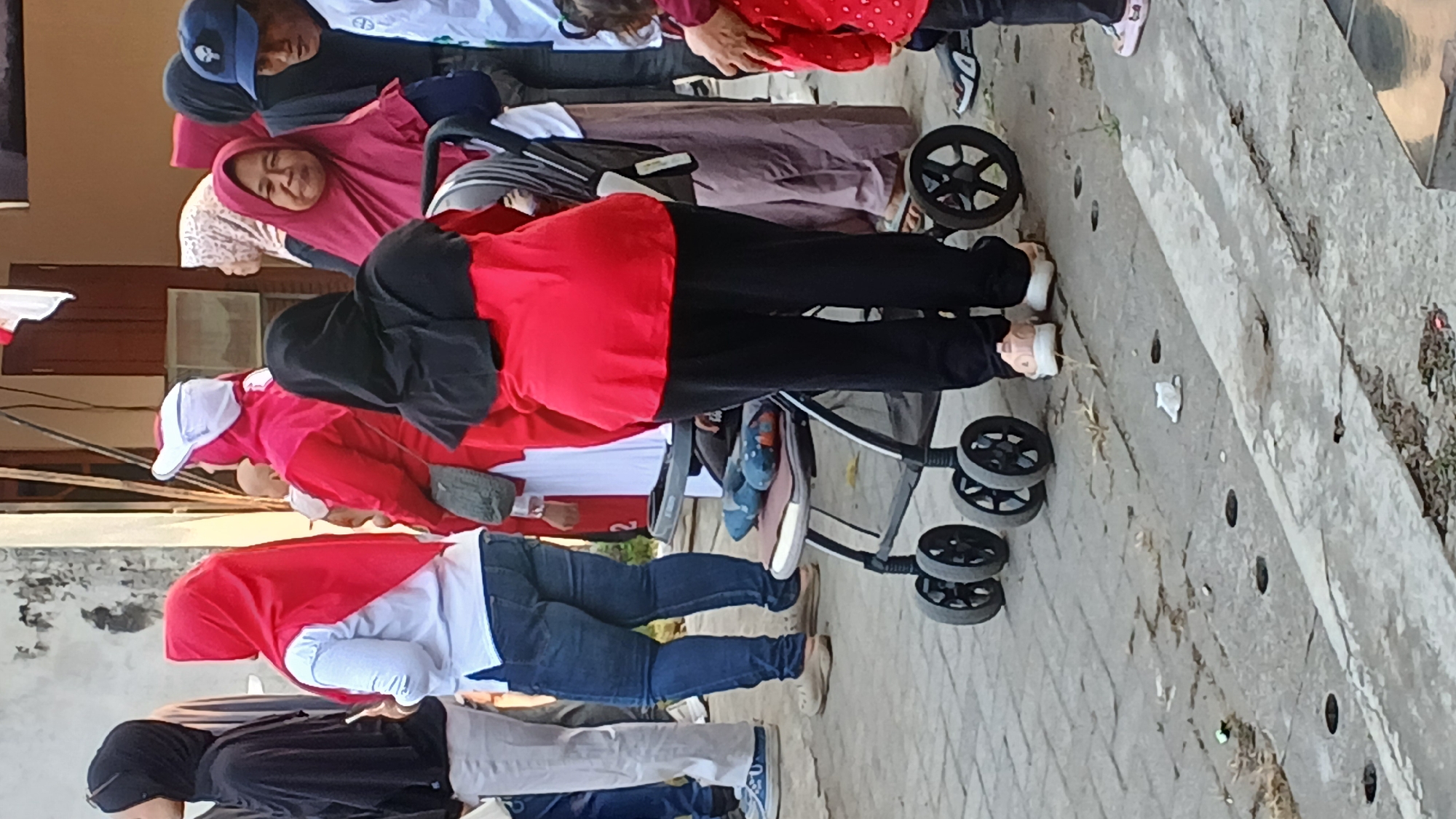 In Mendut, Indonesia, several individuals are gathered outdoors, possibly for an event. A woman wearing a red hijab and a red top is the central figure, facing away from the camera. She is pushing a stroller with black wheels. To her left, a woman in a red hijab and white shirt is visible, and further left, a person in a black hijab and light-colored pants. To the right of the central figure, another person in a pink hijab is looking forward. Behind them, other people in various attire, including a man wearing a blue baseball cap, are present. The ground is paved with concrete blocks, and some dry leaves and debris are scattered on the surface. The scene appears to be a public space, possibly near a building, with a hint of a red and white flag in the background.