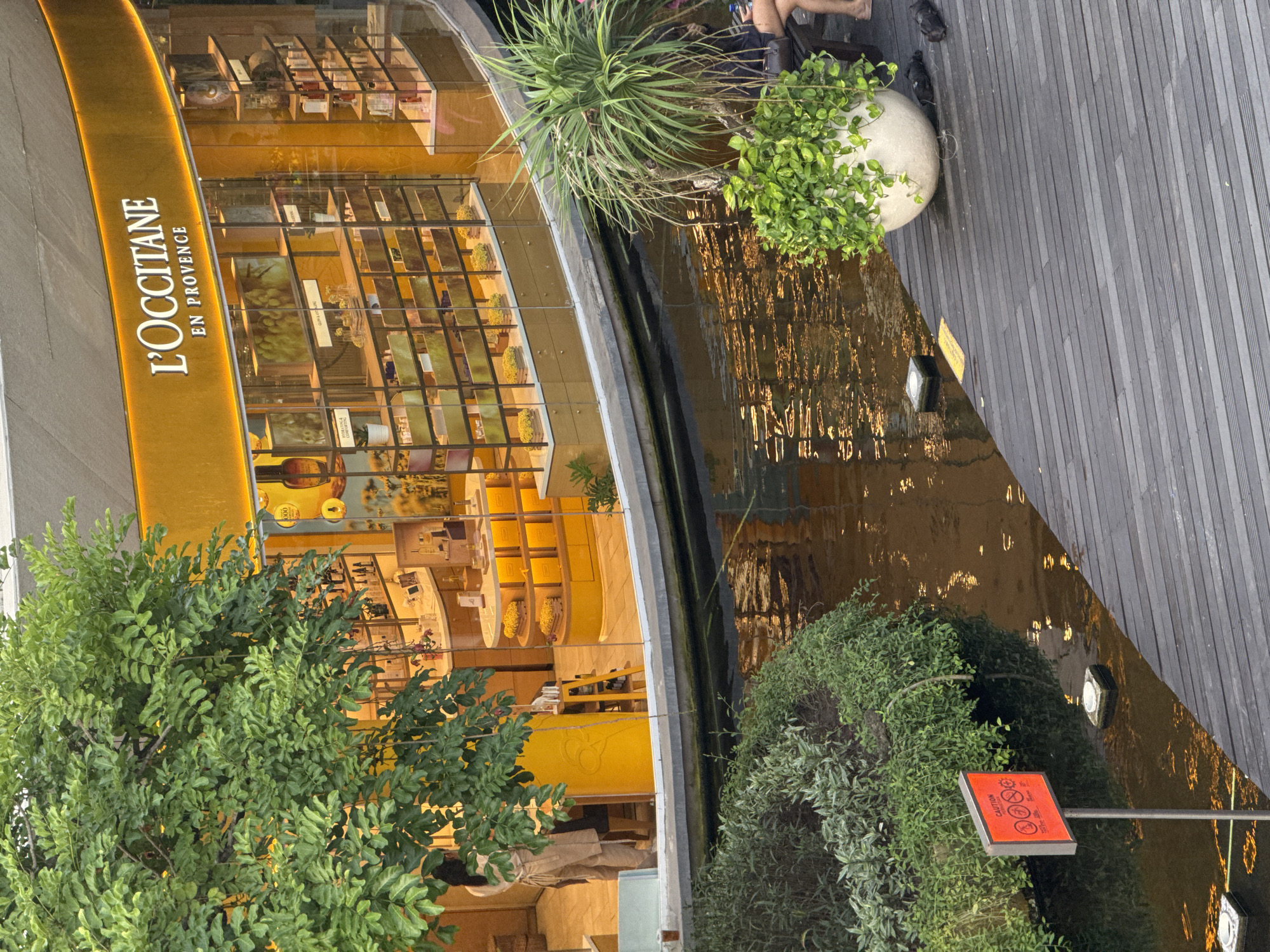 The image shows a partial view of L'Occitane en Provence shop in Kuta, Indonesia. The shop features a curved glass front with displayed products. A water feature runs alongside the shop, reflecting light. Next to the water feature, there's a wooden deck and plant pots with greenery. A caution sign is visible near the water's edge, surrounded by bushes. Some people are partially visible. The surrounding environment includes other plants and buildings.