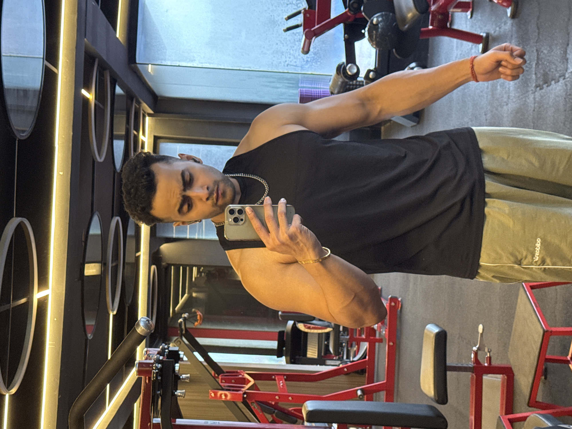 A young adult male is taking a mirror selfie in a gym. He is wearing a black tank top and light green athletic shorts. He has a silver chain necklace and a gold bracelet on his left wrist. A red string bracelet is visible on his right wrist. The gym appears to be modern and well-equipped with various weight machines and equipment, including red-framed weight machines and black benches. The lighting is bright, with circular light fixtures visible on the ceiling. The floor is dark and textured, consistent with gym flooring. The background shows gym equipment and a large window with frosted glass. The man's expression is serious as he focuses on his phone. The image does not contain any information that would allow for a specific location within Kolkata, India to be identified.