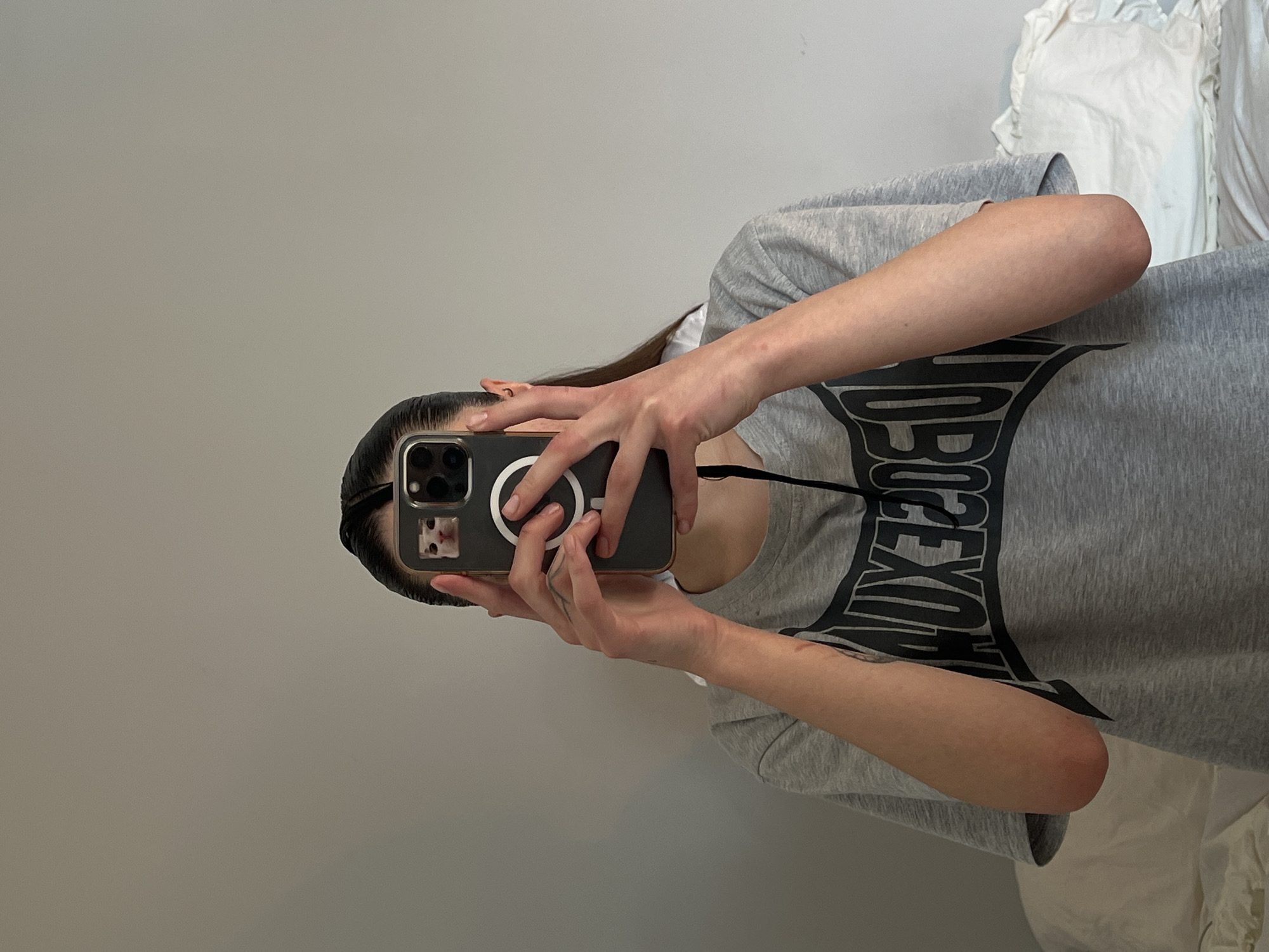 A person stands indoors, facing a light gray wall, and taking a selfie in a mirror. The person holds an iPhone encased in a black case with a MagSafe charging ring. The phone appears to have a cat photo in the corner, which is a sticker. The person wears a gray t-shirt with black text. A white garment is visible in the background. It is not possible to determine the location in Ternopil, Ukraine from the given image.
