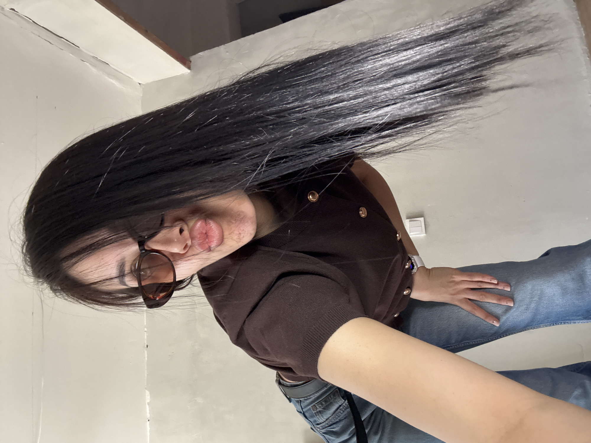 A woman is posing for a selfie indoors. She has long, dark hair that is spread out. She is wearing brown sunglasses, a brown cardigan with gold buttons, and blue jeans. She is making a kissy face. The background includes a white wall and ceiling. The location is not evident within the city of Votkinsk, Russia, as it is not visible in the image.