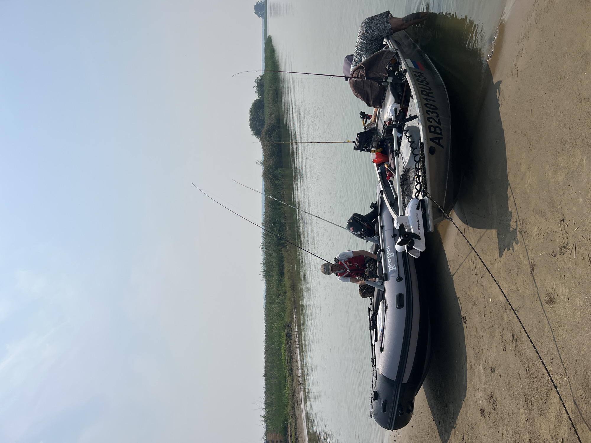 Two inflatable and aluminum boats are partially beached on a sandy shore, with their bows facing the water. The inflatable boat, on the left, is grey and black with the brand name "River Boats" visible. A person wearing a white shirt and a red life vest is seated in this boat, holding a fishing rod. The aluminum boat, on the right, is larger and has the registration number "AB2301RUS54" with the Russian flag emblem painted on its side. Another person, wearing a camouflage-patterned shirt and shorts, is partially submerged in the water, appearing to be adjusting something on the right side of the aluminum boat. Several fishing rods are visible in both boats, and the water is calm with a faint reflection of the sky. The background features a calm body of water and a vegetated shoreline with reeds and scattered trees, under a hazy, light blue sky. The overall scene suggests a fishing excursion near Novopichugovo, Russia.