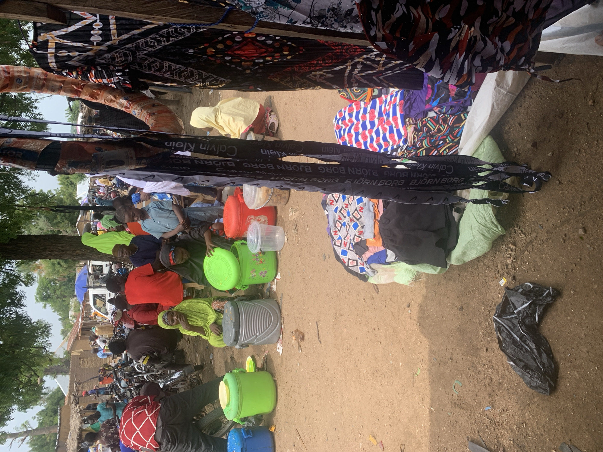 This media file captures a vibrant outdoor market scene in Nigeria. The foreground is dominated by colorful fabrics, some appearing to be clothing or textiles, hanging from an overhead structure, creating a canopy. Below, a diverse group of people are seen engaged in various activities. Some are seated, possibly vendors or shoppers, while others appear to be standing or moving through the market. Several motorcycles are parked, suggesting transportation is a key element of the setting.
Various objects are scattered throughout the scene, including bright green plastic buckets, grey containers, and what appear to be water jugs or storage bins. A black plastic bag lies on the ground in the lower right corner, hinting at the presence of waste or discarded items. In the background, simple structures, likely market stalls or homes, are visible, along with trees and vegetation, indicating a rural or semi-urban environment. A white truck with an umbrella on top adds to the bustling atmosphere.
The lighting suggests it might be daytime, possibly late morning or early afternoon, with the sun casting shadows. The overall mood appears to be one of daily life and commerce, with people actively participating in the market.
Visible text in the image includes brand names such as "Calvin Klein" and "BJÖRN BORG" repeated on fabric straps or ribbons, possibly part of the merchandise being sold. The scene depicts a typical day at a Nigerian market, alive with activity, trade, and community interaction.