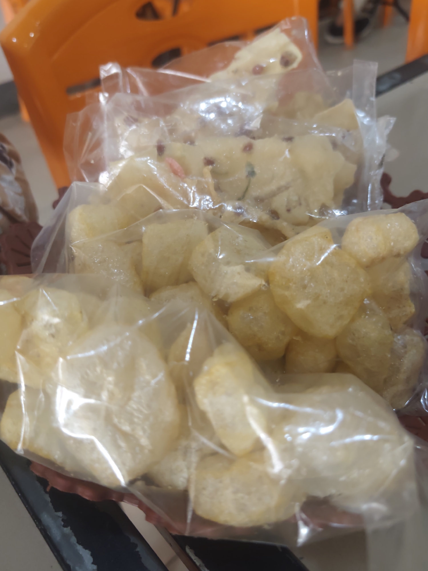 The image shows a close-up of several plastic bags filled with what appear to be fried crackers or chips. The items are light yellow and irregular in shape, with some visible texture. The bags are transparent, allowing the contents to be seen. The items are contained within a shallow, dark red bowl or basket. In the background, an out-of-focus orange plastic chair is visible. The scene appears to be indoors, possibly on a table, with a blurry floor or surface in the foreground. There are no people or distinct actions depicted in the image, and the location in Pematang Reba, Indonesia, cannot be determined from the visual information provided.