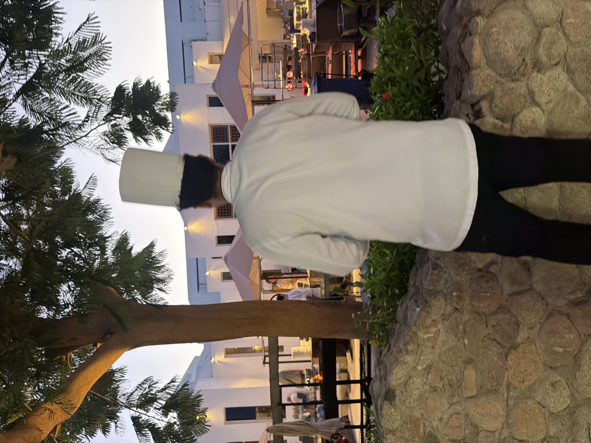 A chef, seen from behind, stands wearing a tall white chef's hat and a white chef's coat. They are looking towards an outdoor buffet setup. In the background, there are white buildings with arched windows and outdoor seating areas covered by umbrellas. The scene appears to be at dusk, with soft lighting illuminating the area. The chef is positioned in front of a stone wall and lush green foliage. The overall setting suggests a hotel or restaurant in Sharm El Sheikh, Egypt, possibly at a resort with an outdoor dining or buffet area.