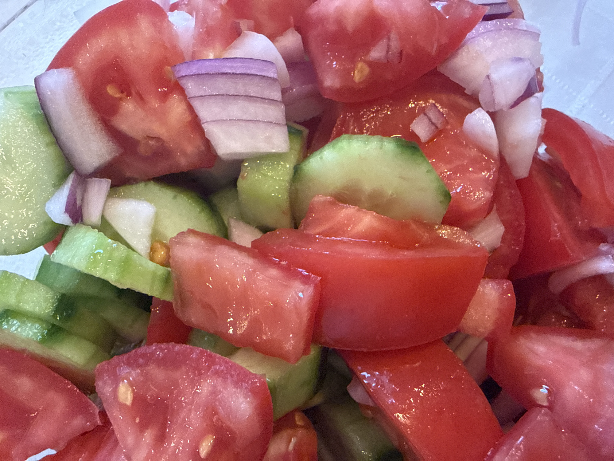 This is a close-up, eye-level, medium shot of a chopped salad. The salad contains chopped tomatoes, sliced cucumbers, and diced red onions. The ingredients are mixed together in a clear bowl, suggesting a casual meal or preparation for one. The lighting appears to be bright, possibly from natural daylight, creating some shine on the wet surfaces of the vegetables. There are no people or text visible in the image. The overall impression is that of fresh, healthy ingredients. The scene is simple and focused entirely on the food. The specific location of Sozopol, Bulgaria, is not visually indicated within the image itself.