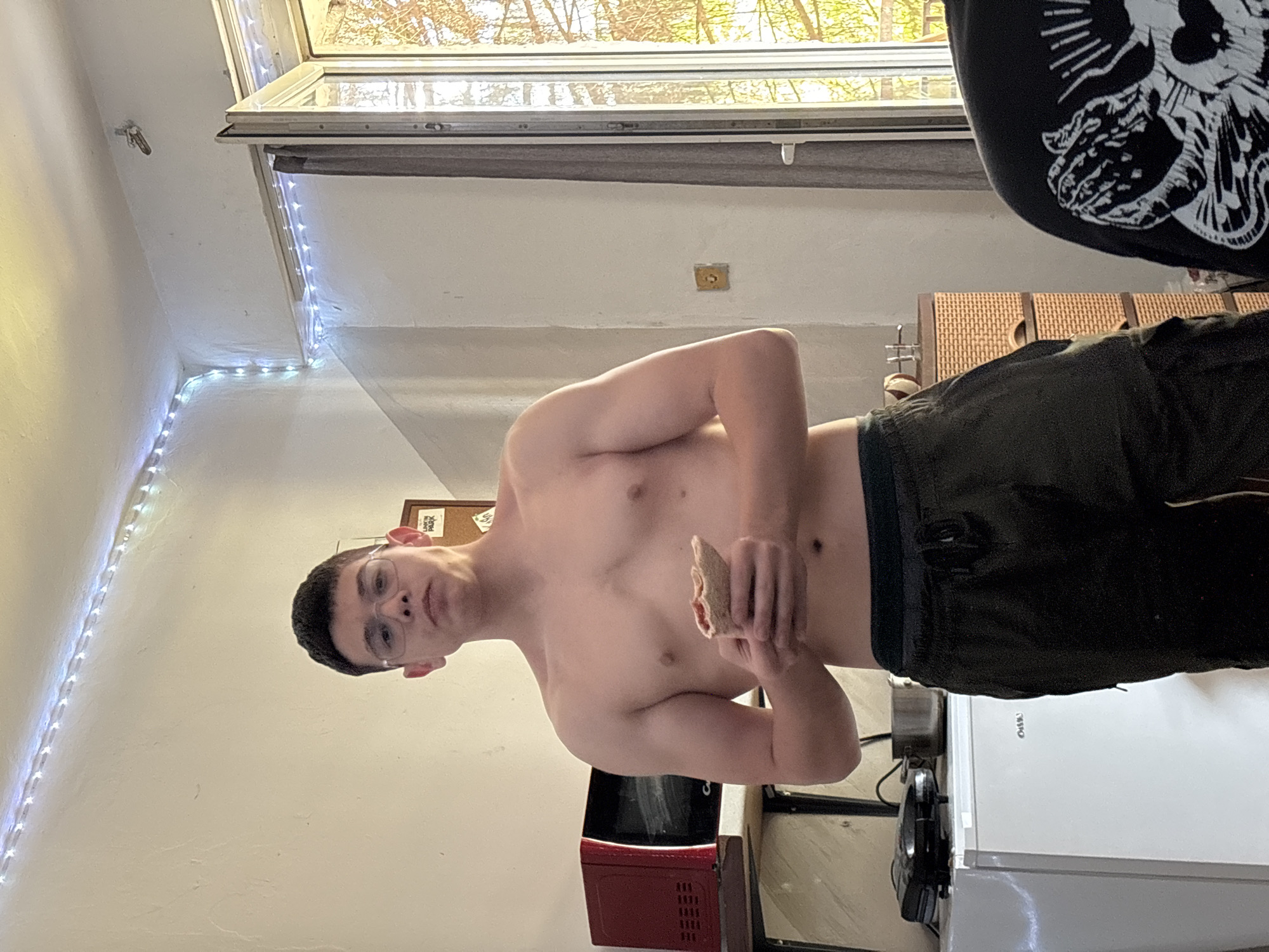 The image captures a young man standing indoors, shirtless and wearing dark athletic shorts, holding a piece of food in his right hand. He looks directly at the camera with a neutral expression. The setting appears to be a home kitchen or a room with kitchen amenities, featuring a red microwave and a white refrigerator visible in the background.
Natural light streams in from a large window located on the right side of the frame (when correctly oriented), revealing trees with green and yellowish leaves outside, suggesting a daytime scene, possibly during spring or early autumn. A dark roller blind is partially drawn across the window. White LED strip lights illuminate the corner of the wall and ceiling on the left side of the room. In the upper right corner, a black garment with an intricate white design, resembling a stylized skull or gothic art, is partially visible.
Further details include a small metallic hook on the wall near the LED lights, and a wooden-framed corkboard partially visible behind the man's head. Visible text includes "LINKIN PARK" on the corkboard and "G-SHOCK" on the refrigerator. The man's casual attire and the act of eating suggest a relaxed, everyday moment within what appears to be a personal living space in Troyan, Bulgaria.