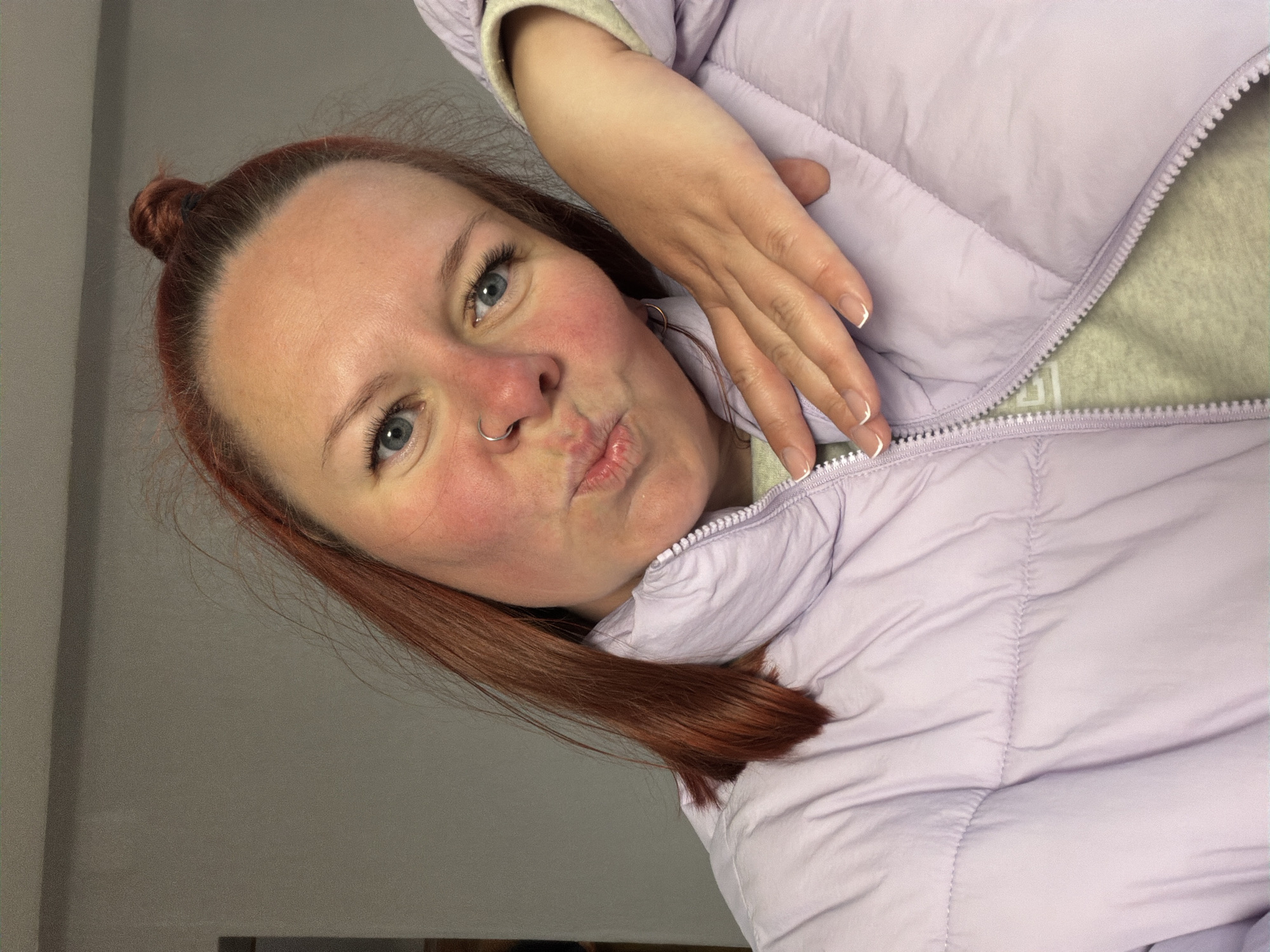 A close-up, rotated portrait features a woman with reddish-brown hair and blue eyes, making a playful "puckered lips" expression. Her hair is pulled back into a bun at the crown of her head, and her fair skin shows rosy cheeks. She wears a silver ring piercing on her left nostril. The woman is dressed in a light purple (lavender) puffer jacket, which appears to have a lighter-colored, possibly fleece, lining visible at the collar and sleeve cuff. Her left hand is visible, resting on her jacket, and her nails are neatly manicured with white French tips. The background is a plain, light grey or beige surface, indicating an indoor setting. On the inside lining of the jacket, near the zipper, a faint, light-colored, and indiscernible logo or geometric pattern is visible. The lighting is even, suggesting an indoor environment.