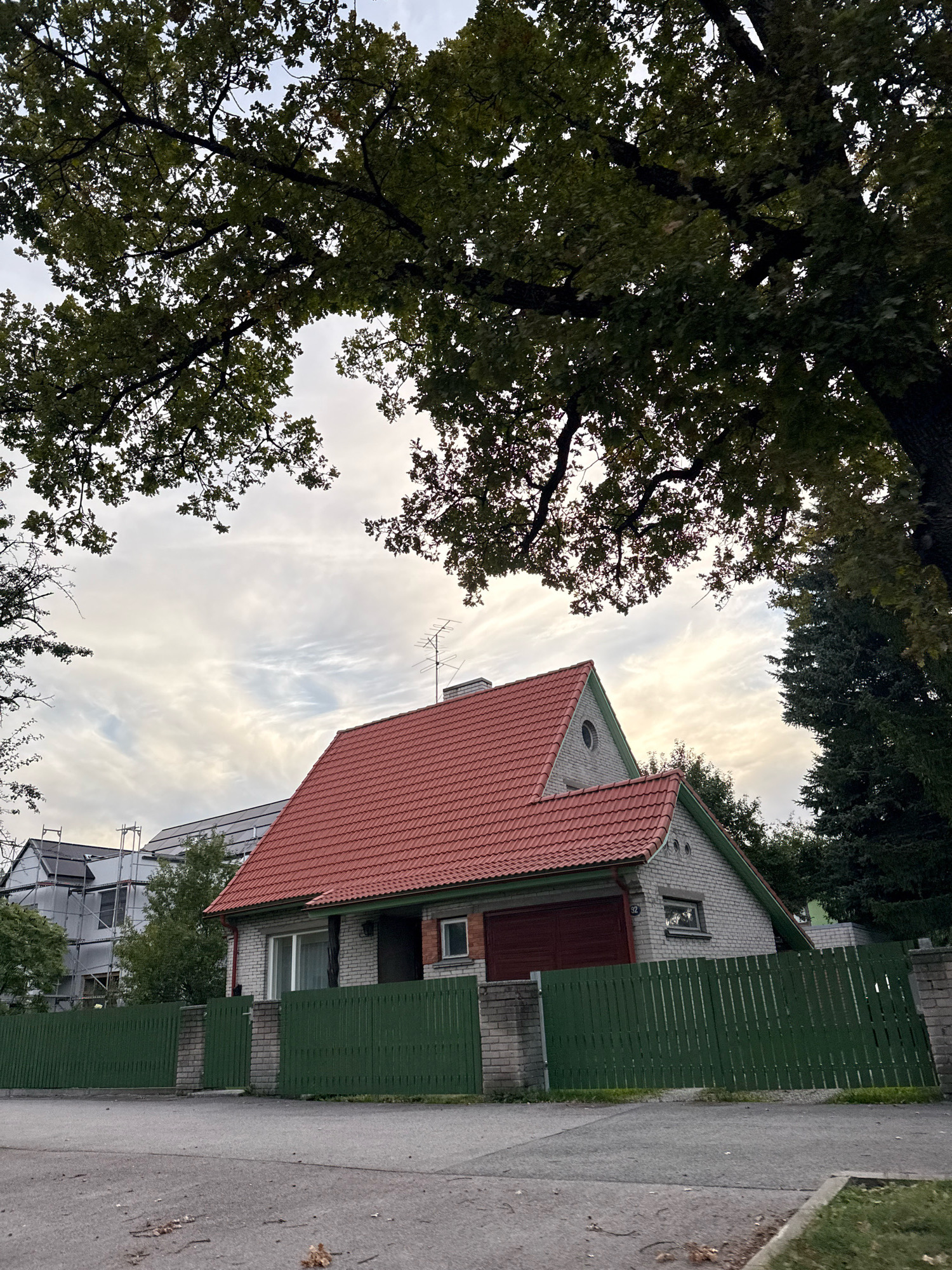 The image depicts a residential street scene in Tallinn, Estonia, under a partly cloudy sky. A light grey brick house with a red tiled roof and a green fence in front is the primary subject. The house has a garage with a dark red door, a white window, and a small, high window on the gable. A chimney and an antenna are visible on the roof. To the left of the house, another building is partially visible, covered in scaffolding and what appear to be solar panels. Large trees with green leaves frame the top of the image, with some branches extending into the frame from the left and right. The street in the foreground is asphalt, with some fallen leaves scattered on it. The overall impression is one of a quiet, suburban residential area.