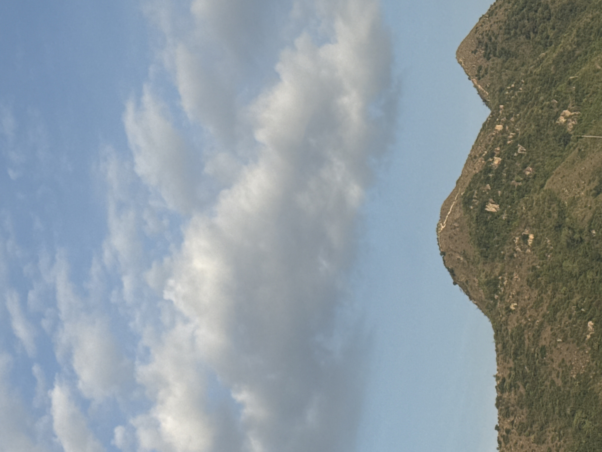 The image shows a panoramic view of two mountains in Bac Nha Trang Ward, Vietnam, under a partly cloudy sky. The mountains are covered in lush green vegetation and appear to be rugged with some rocky outcrops. A winding path is visible on the slopes of the left mountain, suggesting it might be a hiking trail. The sky is a clear blue with scattered white clouds, casting soft shadows on the landscape. The overall scene evokes a sense of natural beauty and tranquility.