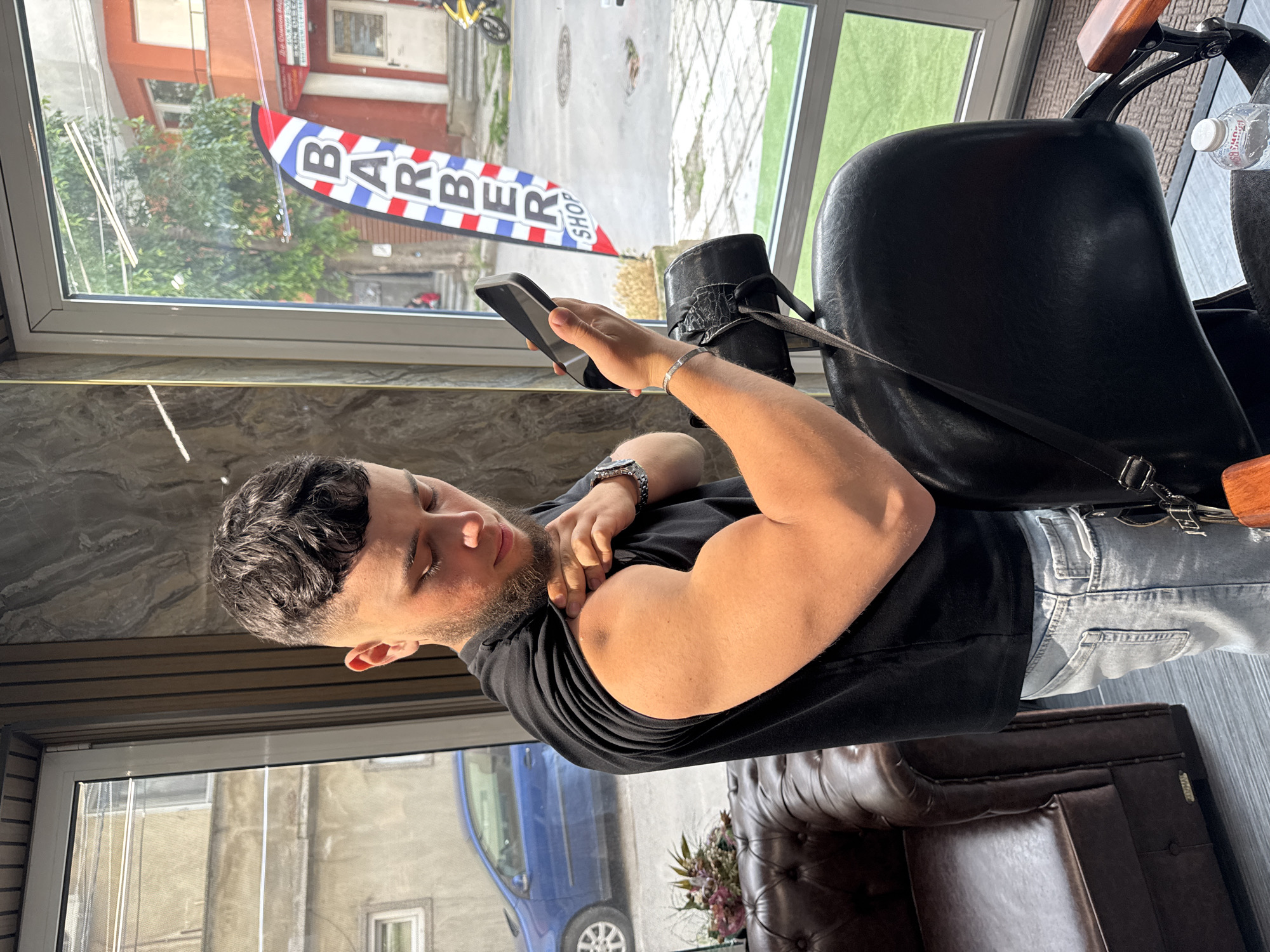 The image depicts a young man inside a barbershop in Varna, Bulgaria, illuminated by abundant natural light from a large window. The man, who has short dark curly hair and a neatly trimmed beard, is seated in a black barber chair. He is dressed in a black t-shirt and light-wash jeans, with a silver watch on his left wrist and a silver bracelet on his right. In his right hand, he holds a black smartphone, while his left hand pulls up the sleeve of his t-shirt, revealing and flexing his right bicep. He appears to be looking down, possibly at his phone or his arm.
Through the large window behind him, an outdoor urban scene unfolds under bright, sunny conditions. A prominent red, white, and blue barber pole-style sign with the text "BARBER SHOP" is clearly visible. Beyond the sign, green trees, various buildings (one with an orange-red facade), a street, a parked blue car, and a yellow bicycle can be seen. Inside the barbershop, a dark grey patterned counter runs below the window, and a section of a brown leather armchair or sofa is visible in the lower right. A clear water bottle rests on the back of the barber chair. The overall mood is casual and well-lit.