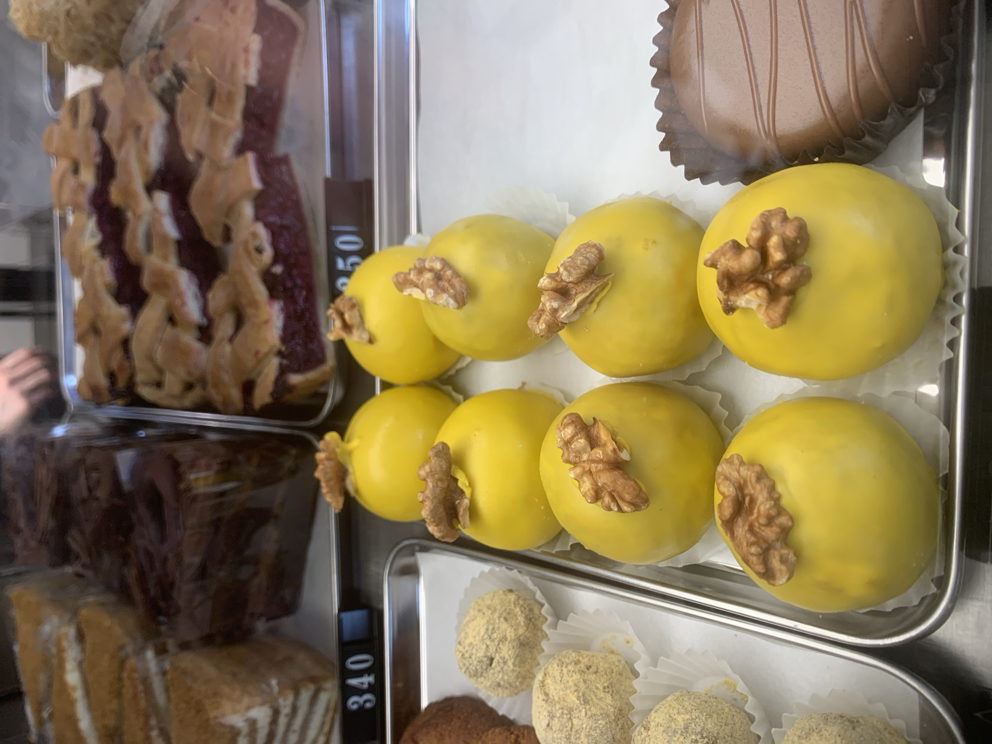 The image presents a display case of pastries in what appears to be a shop in Izhevsk, Russia. The pastries are arranged on silver trays inside a glass display case. The upper tray holds a row of six yellow, round pastries, each topped with a walnut. Above them, in the same tray, is a chocolate pastry in a fluted cup. A tray containing small, round, brownish pastries is below the yellow pastries.
To the left of the trays, through the glass, are additional pastries or cake slices, which appear to have fruit layers and cream. Price tags on the divider are visible: "340" and "550". A sliver of someone's hand is visible through the left side of the display case. The environment is likely an interior shop setting, with a focus on the array of pastries available for purchase.