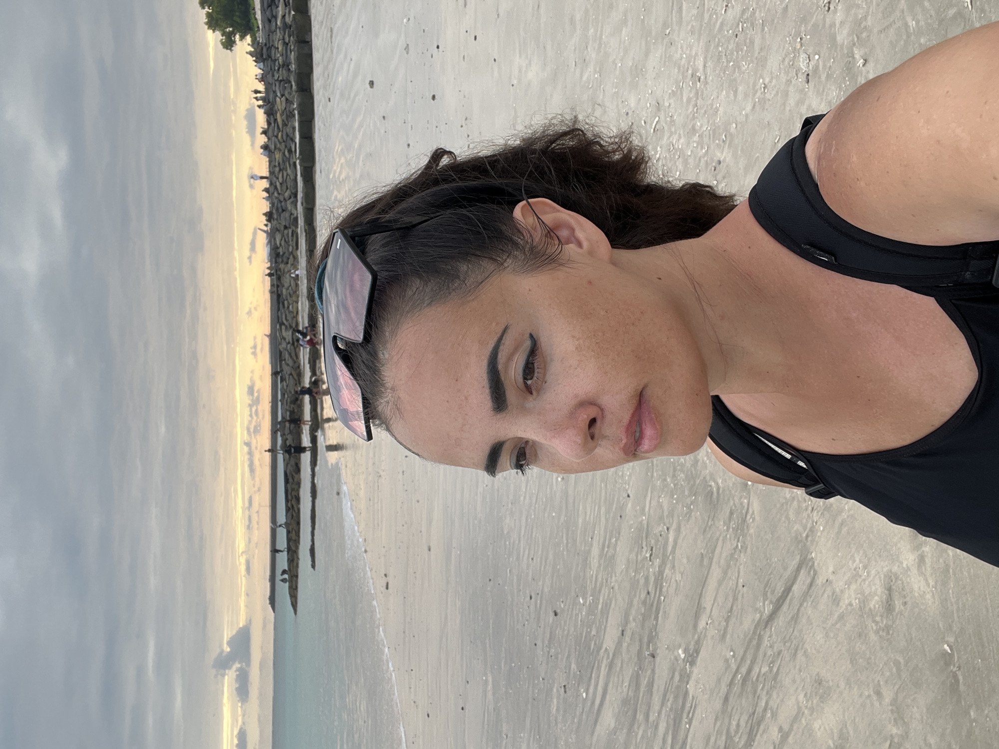 An image taken at Kuta Beach in Bali, Indonesia, shows a woman with sunglasses perched on her head, looking to the left. She is wearing a black tank top and a black backpack. The beach is sandy, with the ocean in the background. The sky is cloudy, with a hint of sunset colors. Several people are visible in the distance, standing on a rocky structure extending into the sea. The overall mood of the image is calm and contemplative.