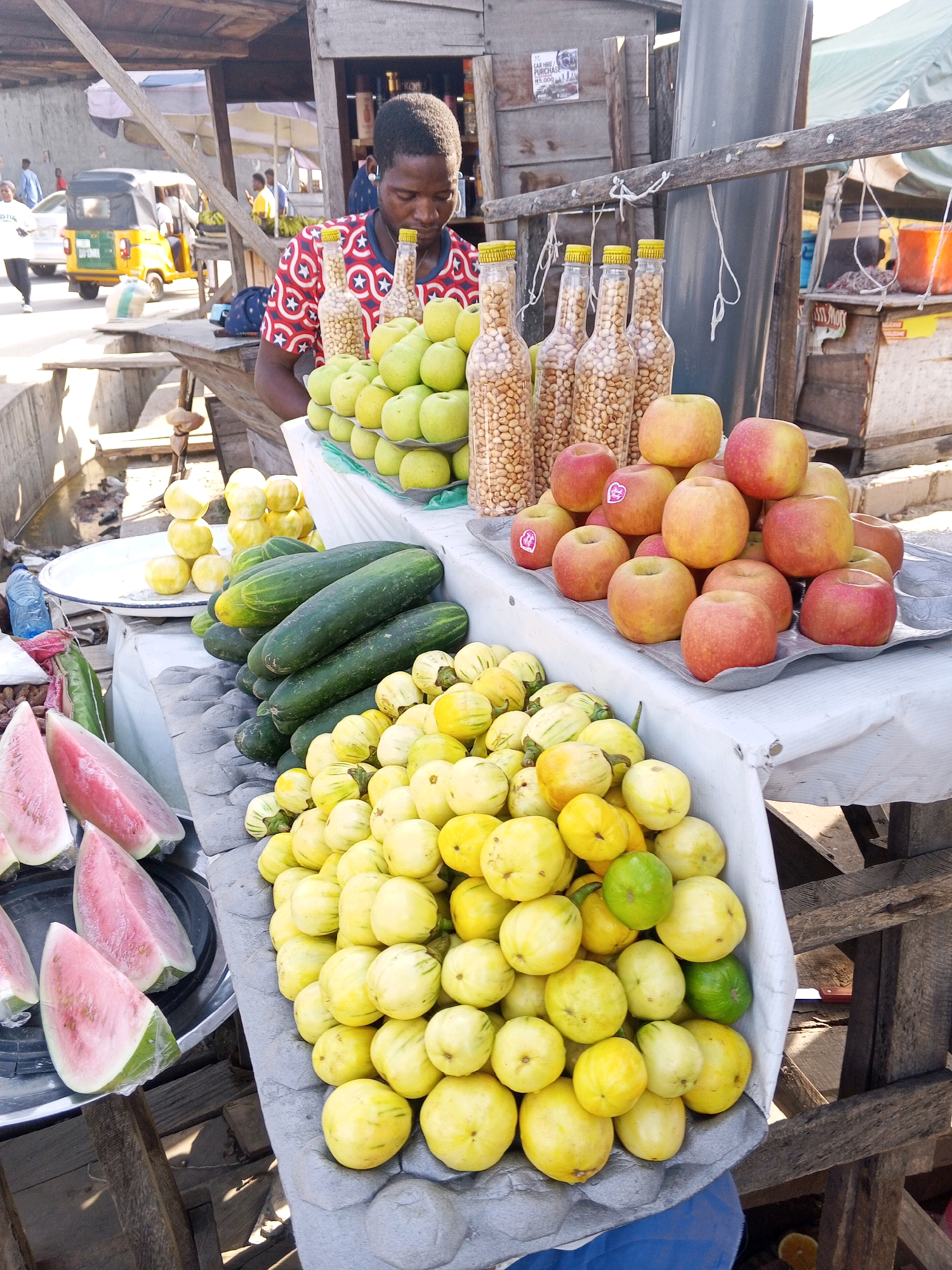 This is a medium-shot, eye-level, slightly tilted photograph of a street market scene in Lokoja, Nigeria. In the foreground, a young man with dark skin and short, black hair, wearing a red and white patterned t-shirt, is attentively arranging a display of produce. He is positioned behind a table covered with a white cloth, which holds various fruits and vegetables.
To the right of the frame, a pile of red and yellow apples is neatly arranged on a gray, textured tray. Behind the apples, four clear plastic bottles filled with what appears to be nuts are standing upright. Further back, a large quantity of small, pale yellow, round fruits, some with striped green skin, are piled high. To the left of these, a collection of dark green cucumbers is stacked.
In the middle ground, on the left side, several slices of watermelon with vibrant pink flesh are visible on a dark plate. A blue plastic water bottle is also present. Behind these items, a white plate holds a stack of round, yellow fruits.
The background depicts the bustling environment of a Nigerian street market. A wooden stall structure with a corrugated metal roof is prominent. In the distance, a yellow and green auto-rickshaw, commonly known as an "okada" or "keke napep," is parked. Other market stalls and people can be vaguely seen in the blurred background, suggesting a busy commercial area. The overall lighting suggests it is daytime, with shadows indicating the sun is overhead or slightly to the side.