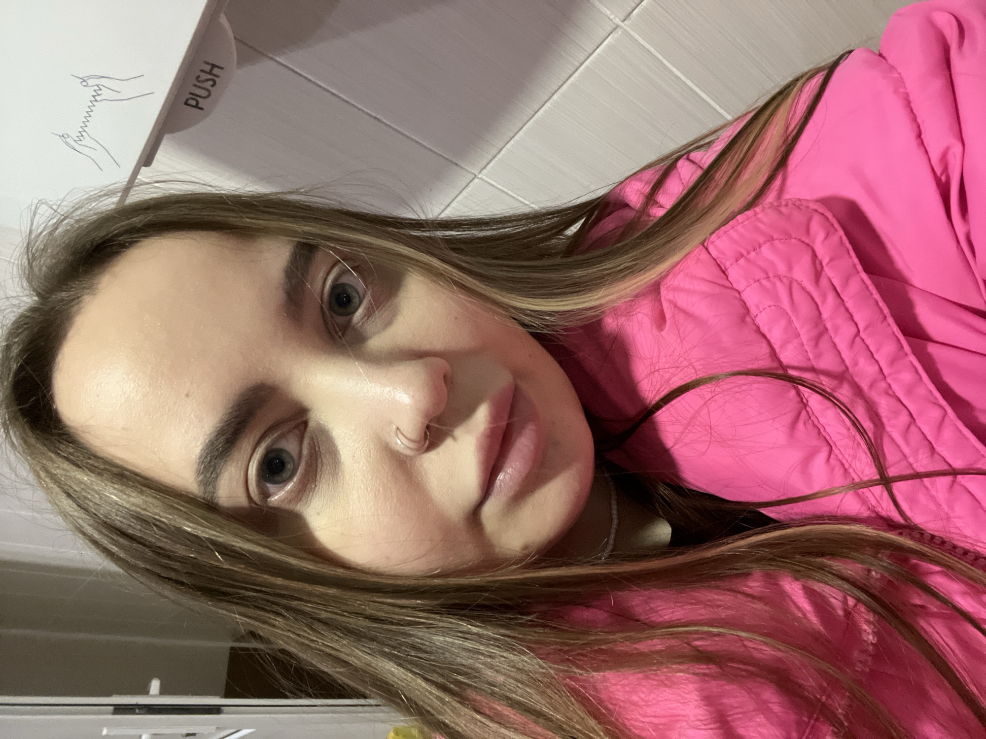 A young woman with long brown hair and a nose ring is facing the camera. She is wearing a bright pink quilted jacket. Behind her is a white wall with tiles and a "PUSH" sign with a graphic of hands. There is no indication of Izhevsk, Russia.