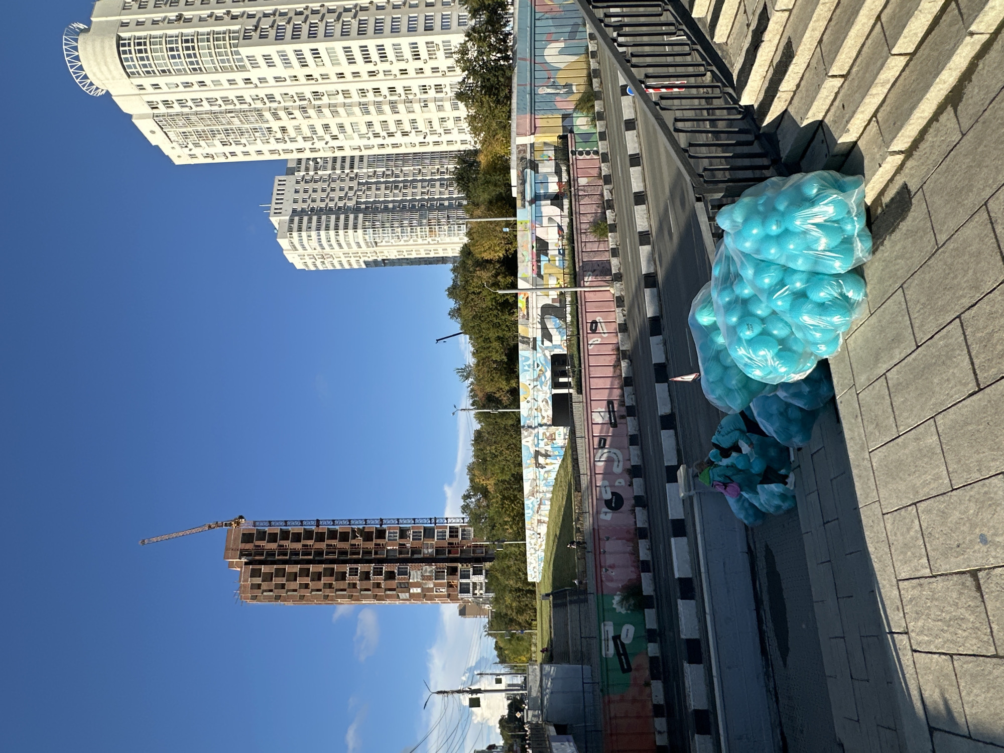 In the city of Unknown, Unknown, a construction site is visible, featuring a tall, unfinished brick building with a crane at its summit. To the right of the construction site, two white high-rise apartment buildings stand completed. In the foreground, several large, transparent plastic bags filled with bright blue balloons are piled on a paved area next to a set of stone stairs. Two individuals, one wearing a green head covering and the other a blue jacket, are interacting with the bags of balloons. Below the paved area, a multi-lane road is visible, bordered by a graffiti-covered wall. The sky is clear and blue.