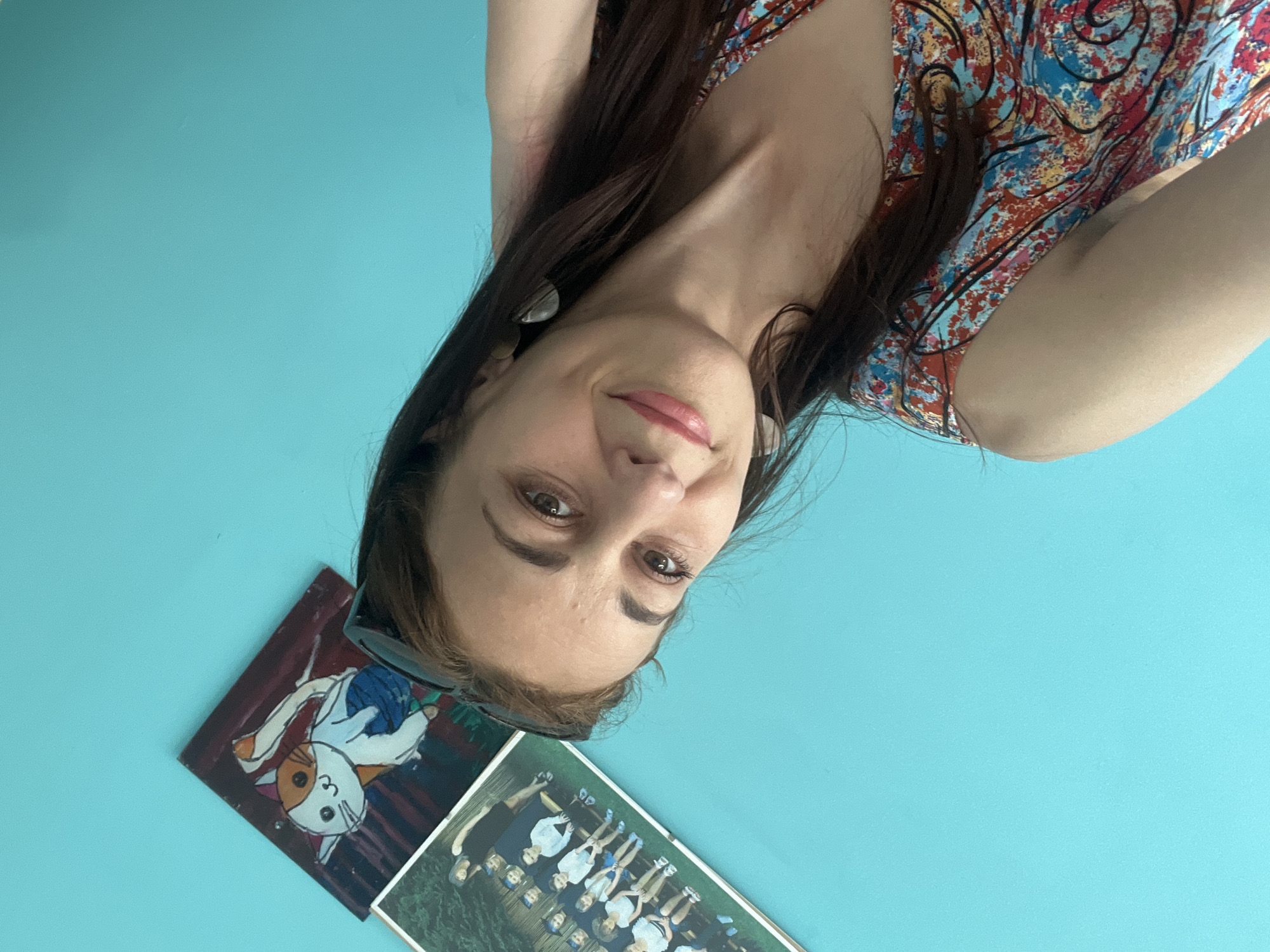The image is a selfie taken by a woman indoors. The woman has long, dark brown hair and is wearing a colorful, patterned dress. She has a light smile and is looking towards the camera. In the background, there are two framed pictures hanging on a bright teal wall. The picture on the left is a painting of a white and orange cat. The picture on the right is a photograph of a group of people, possibly children, in uniform. The image is taken from a slightly downward angle, with the woman's face being the primary focus. The lighting appears to be natural and diffused. The overall mood of the image is casual and personal.