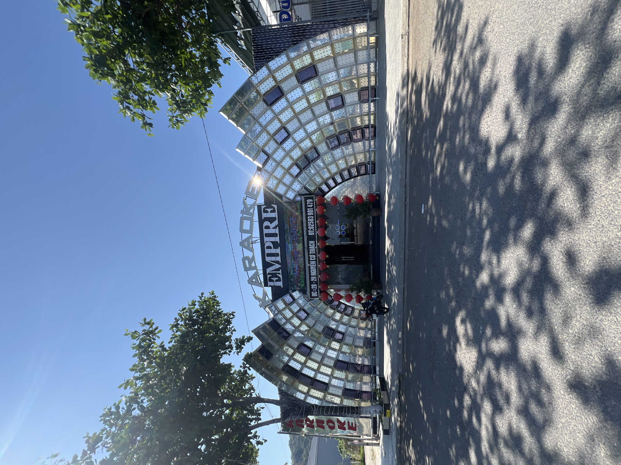 The image displays the exterior of the "KARAOKE EMPIRE" establishment, located at 28-29 Nguyễn Cơ Thạch street in Nha Trang, Vietnam. The building's prominent facade features a large, arched structure composed of numerous rectangular glass blocks, some clear and some tinted dark, creating a reflective, wave-like design. Above the dark glass entrance doors, a large sign reads "KARAOKE EMPIRE" in white letters, with the address and phone number "ĐT: 02583 901 479" displayed beneath. Red spherical lanterns hang strung across the entrance. A single black motorcycle is parked on the sidewalk directly in front of the entrance. Two potted plants are visible near the doors. The scene is illuminated by bright sunlight under a clear blue sky, with a visible sun flare near the top center. Two large trees with green foliage frame the building, casting distinct, dappled shadows across the asphalt street in the foreground. A tree-covered hill is visible in the background to the left of the building. No individuals, actions, or interactions are depicted.