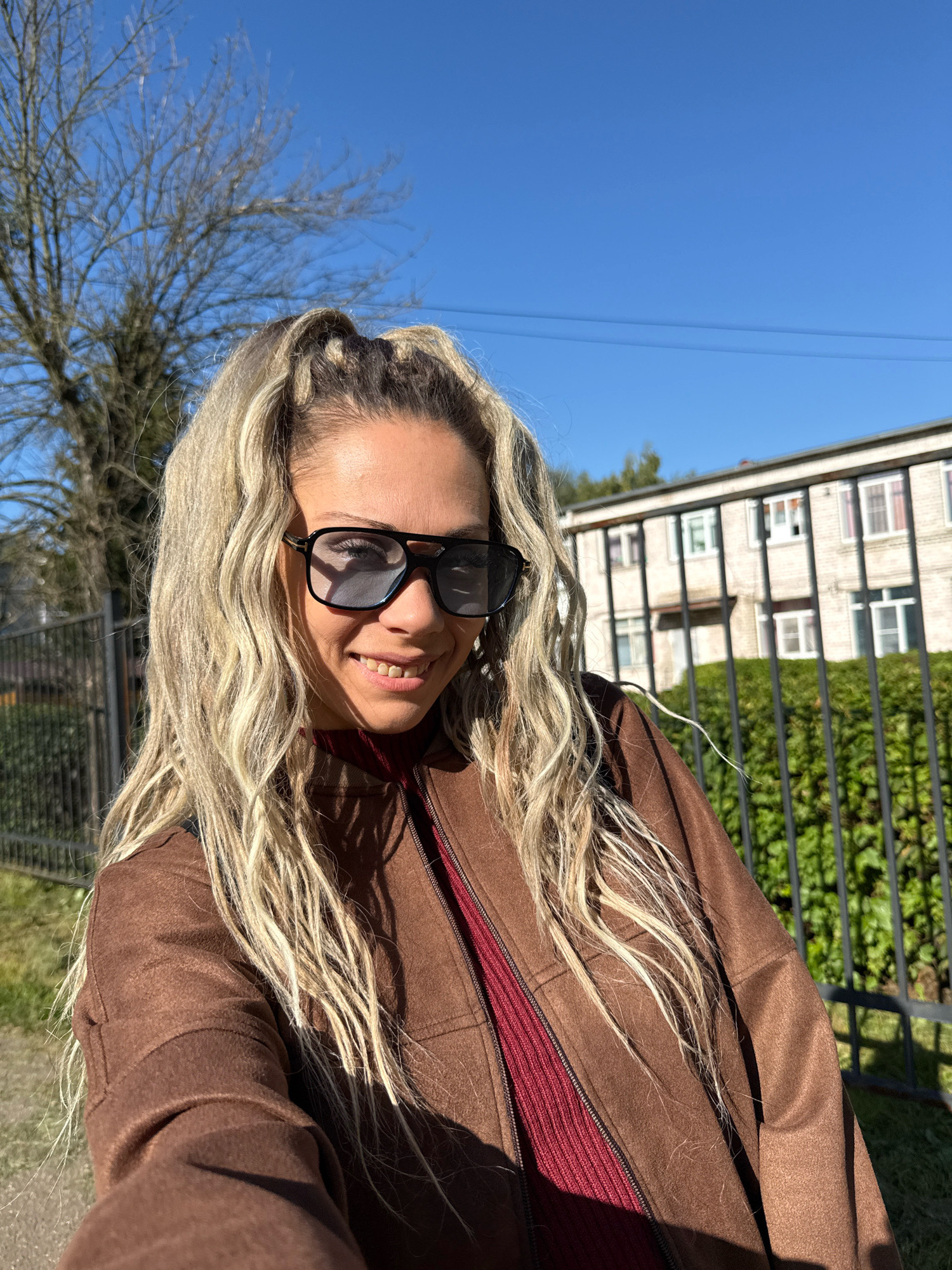 In Rusanovo, Russia, a woman with blonde, wavy hair pulled into a high ponytail stands outdoors in front of a building. She is wearing dark sunglasses, a maroon ribbed turtleneck, and a brown jacket. The background shows a black metal fence, green bushes, a bare tree, and a light-colored building with multiple windows. The sky is clear and blue.
